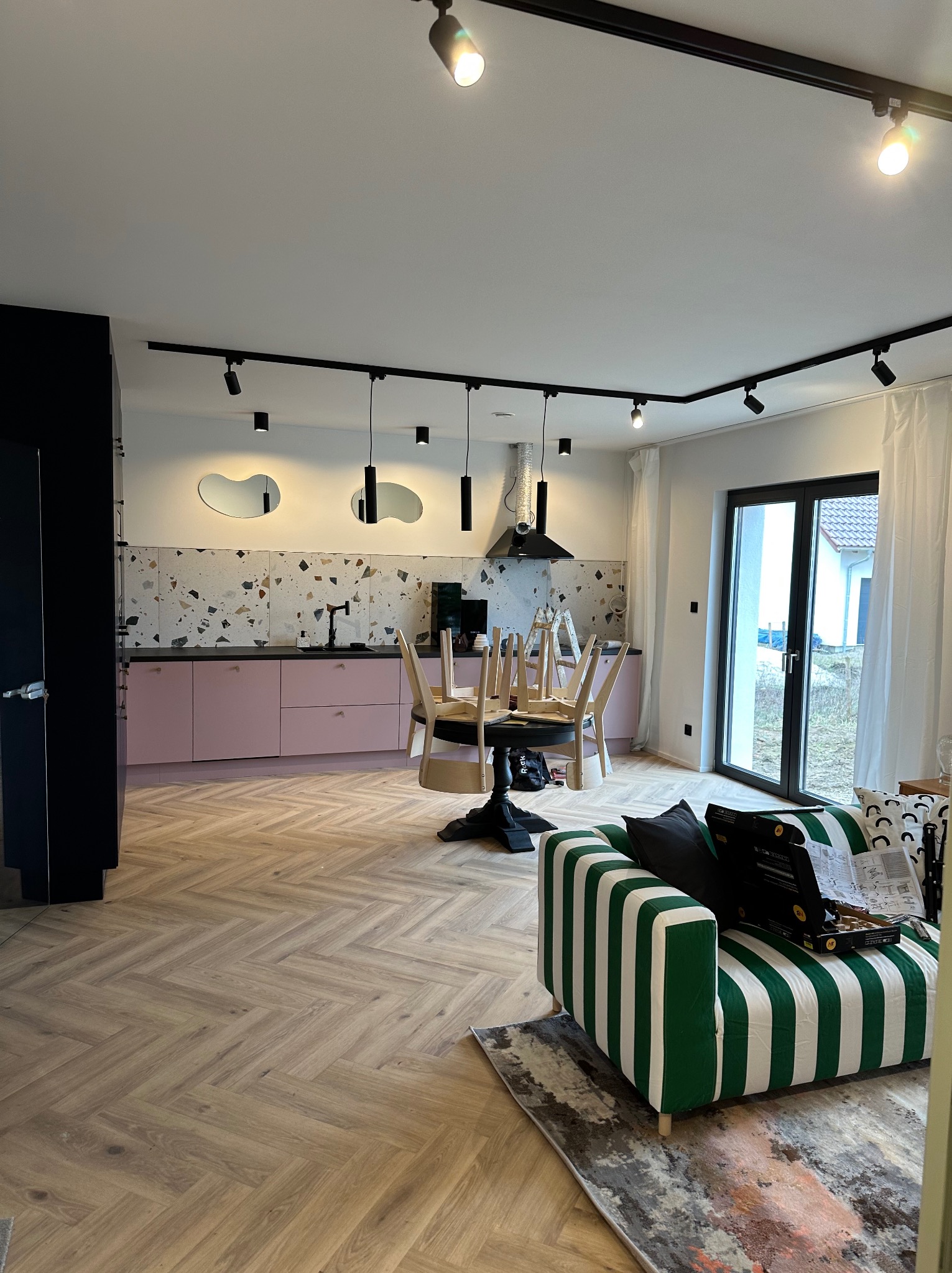 Nowoczesna kuchnia w trakcie remontu: różowe szafki, blat w terrazzo, oświetlenie szynowe, krzesła na stole, drabina i zielono-biała sofa w salonie.