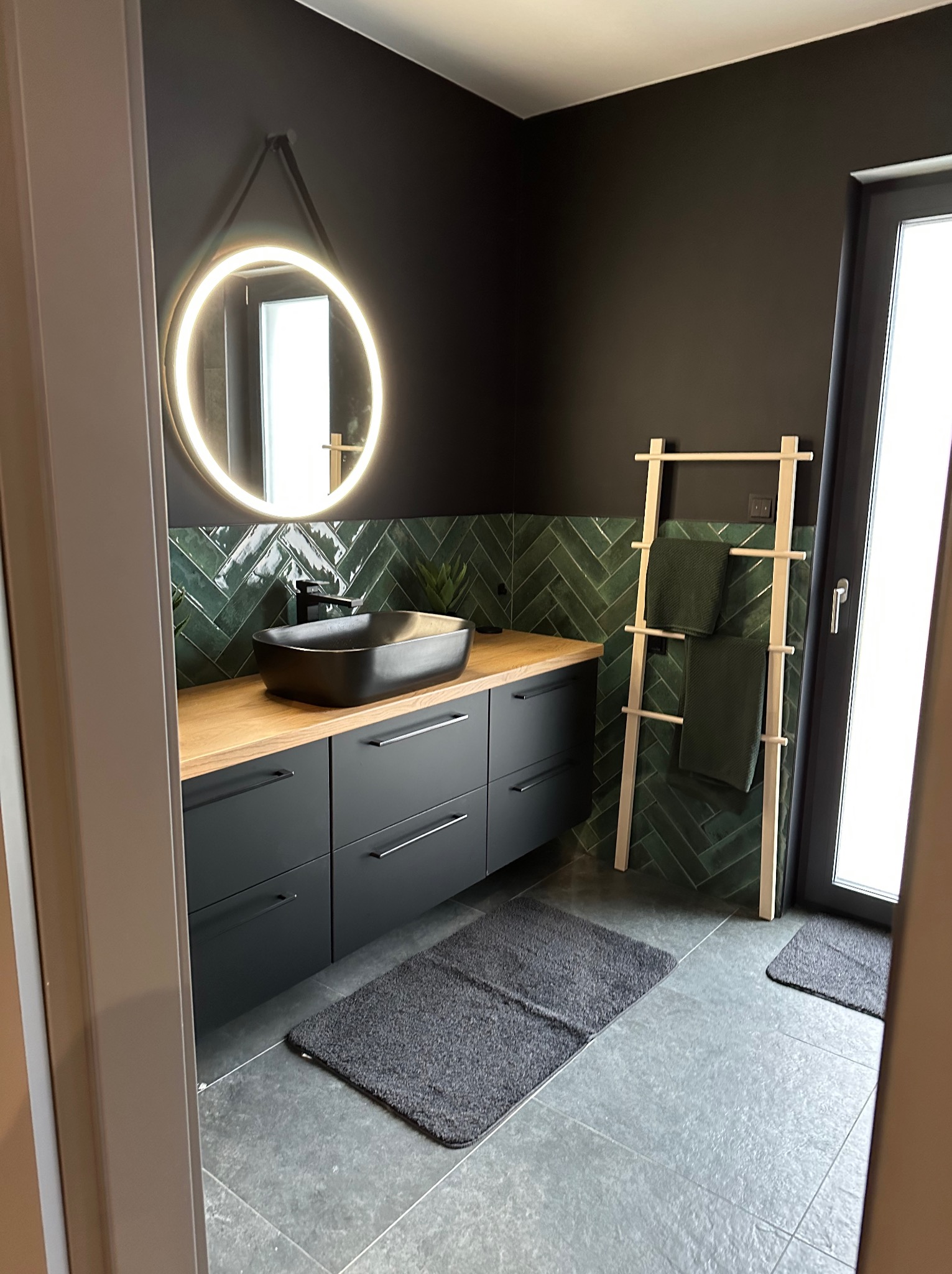 Nowoczesna łazienka z ciemnymi ścianami, drewnianym blatem i umywalką nablatową, podświetlane okrągłe lustro, zielone płytki ułożone w jodełkę na ścianie, drewniany wieszak na ręczniki