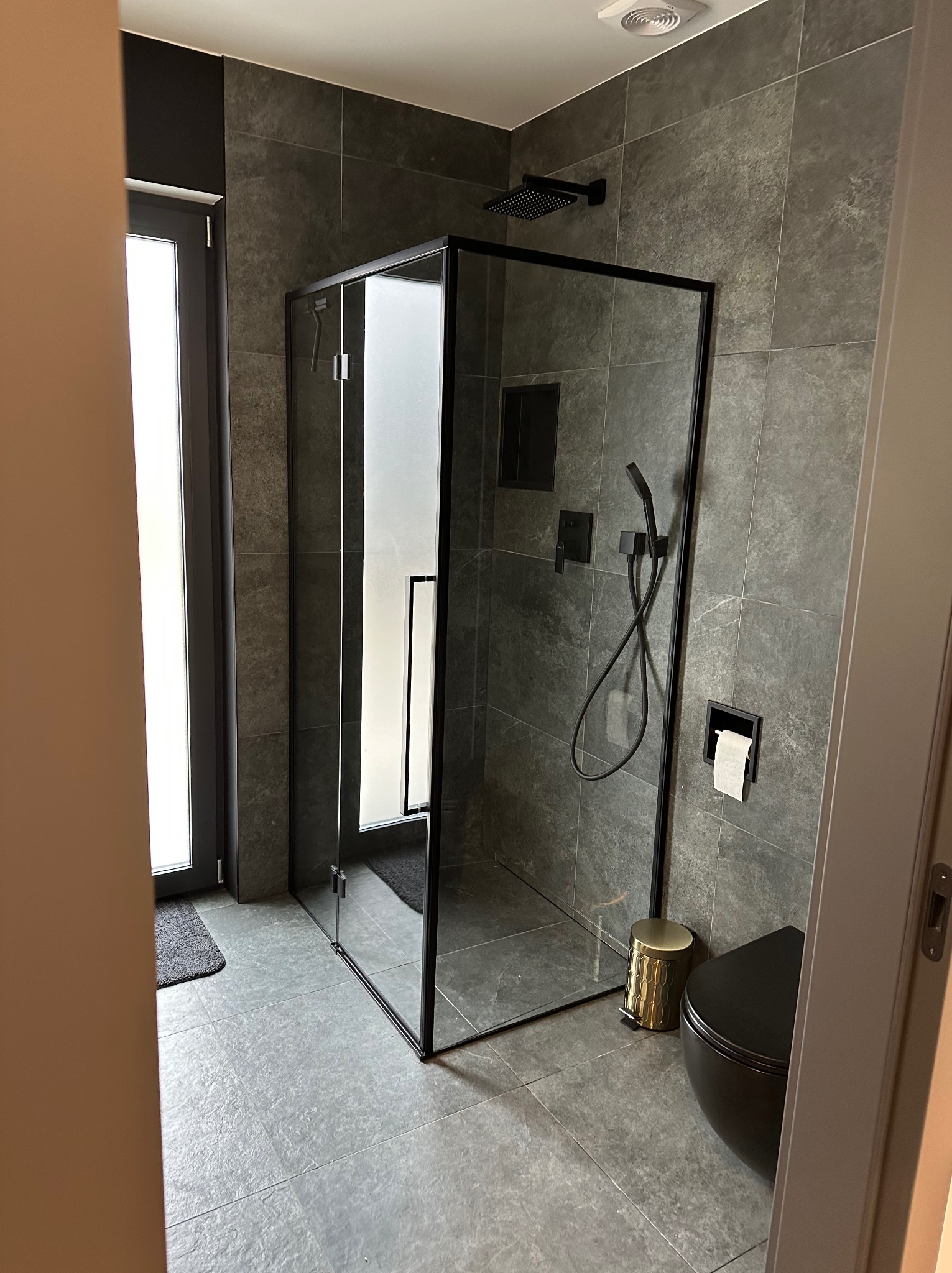 Nowoczesna łazienka z szarymi płytkami imitującymi kamień, kabiną prysznicową z czarnymi ramami, czarną toaletą wiszącą i złotym koszem na śmieci.