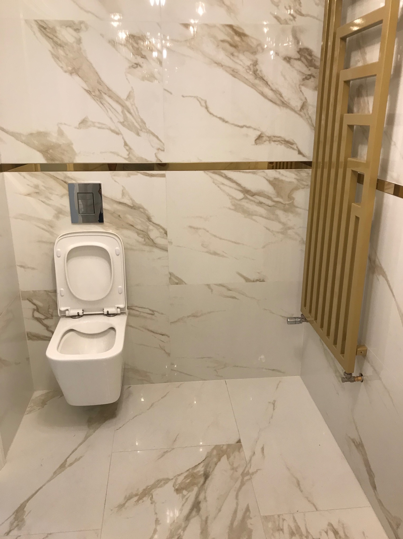 Nowoczesna toaleta z otwartą klapą, wykończona płytkami imitującymi marmur z złotymi akcentami i ozdobnym grzejnikiem drabinkowym w kolorze złota.