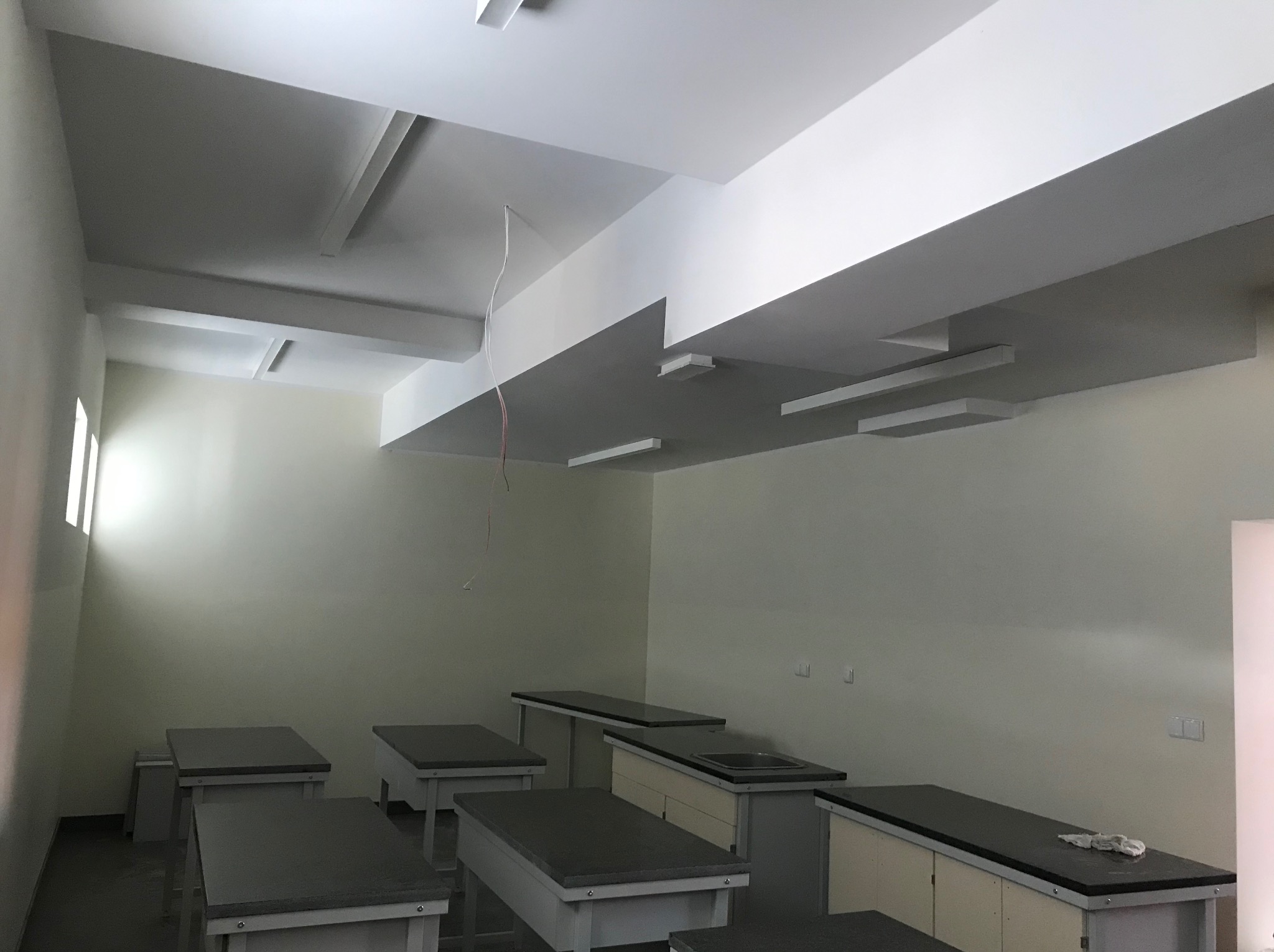Wnętrze laboratorium lub pracowni po remoncie, z widocznymi pomalowanymi ścianami, nowym oświetleniem sufitowym i zainstalowanymi blatami roboczymi z umywalką, częściowo zakrytymi folią ochronną.