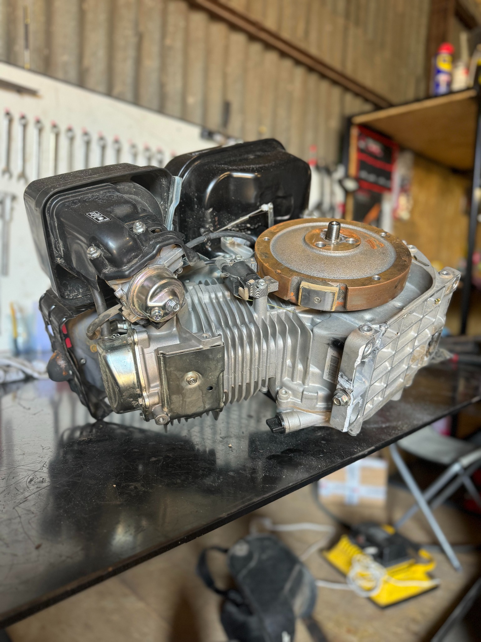 Silnik spalinowy czterosuwowy rozebrany na stole warsztatowym, widoczne żebra chłodzące cylindra, gaźnik i koło zamachowe, narzędzia w tle.
