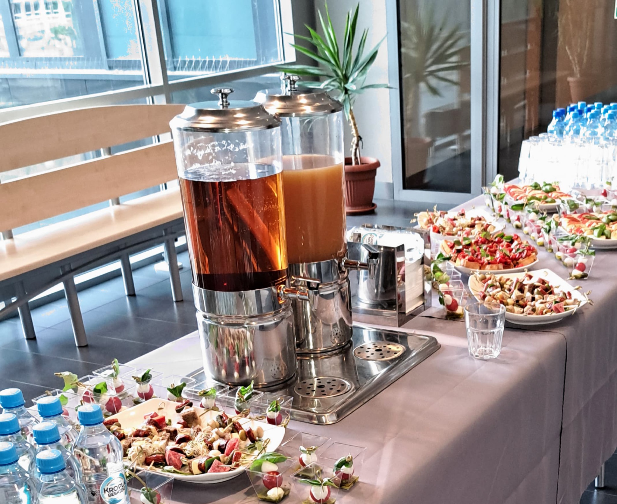 Stół z przekąskami i napojami w dyspenserach, przygotowany na firmowe spotkanie w nowoczesnym biurze z dużymi oknami i roślinami doniczkowymi w tle.