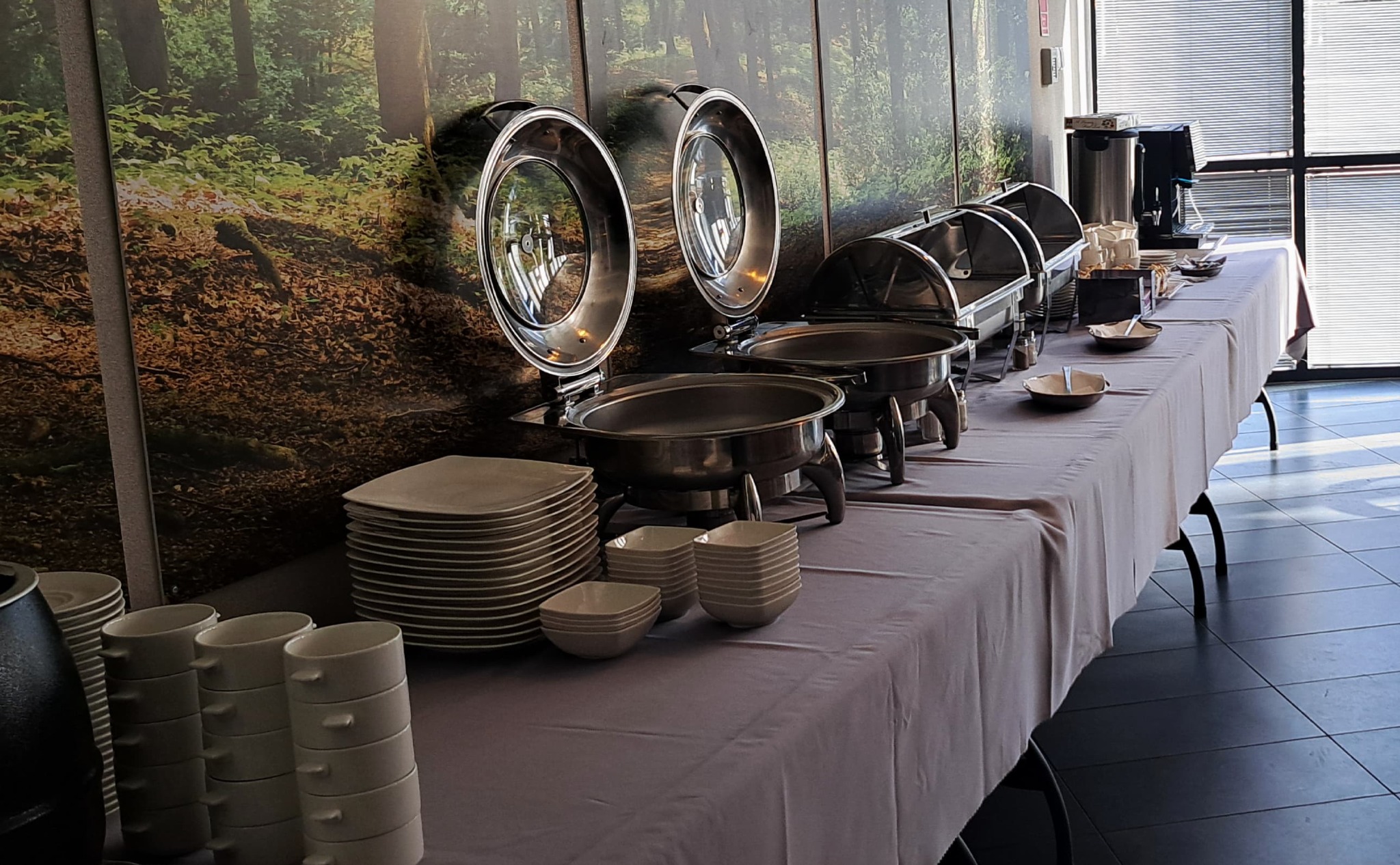 Bufet z podgrzewaczami, stosem talerzy i filiżanek na stole z szarym obrusem, w tle fototapeta z motywem leśnym i okno z żaluzjami.