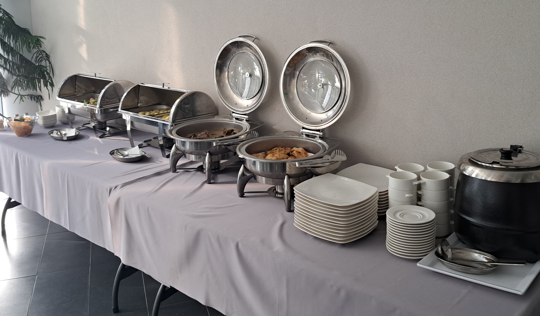 Stół z szarym obrusem, na którym znajdują się podgrzewacze ze stali nierdzewnej z jedzeniem, stosy białych talerzy i filiżanek oraz czarny termos na zupę.