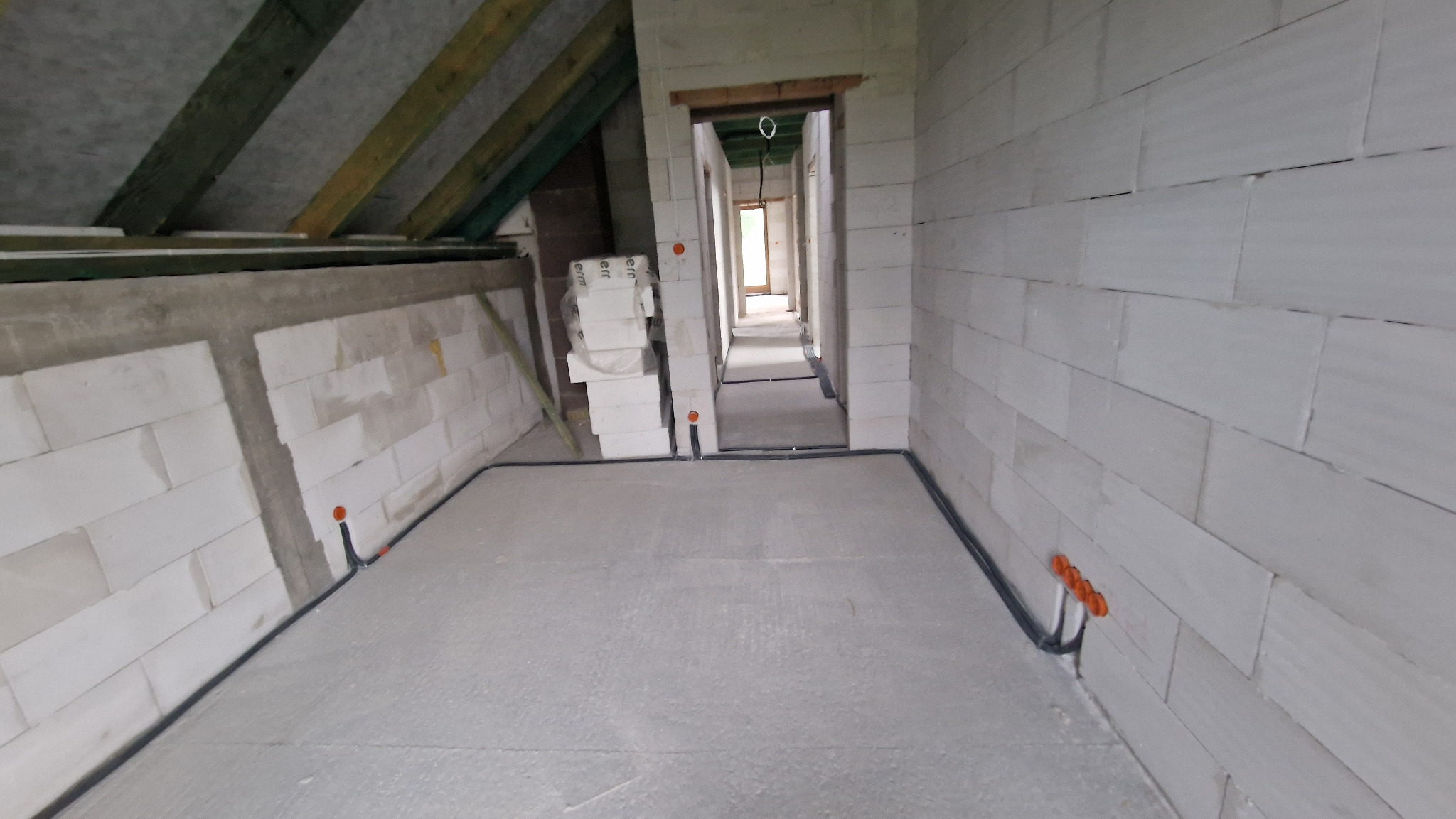 Wnętrze poddasza w trakcie budowy, widoczne ściany z białych bloczków, strop z drewnianych belek, czarne rury instalacji elektrycznej prowadzone wzdłuż ścian, pomarańczowe puszki elektryczne.