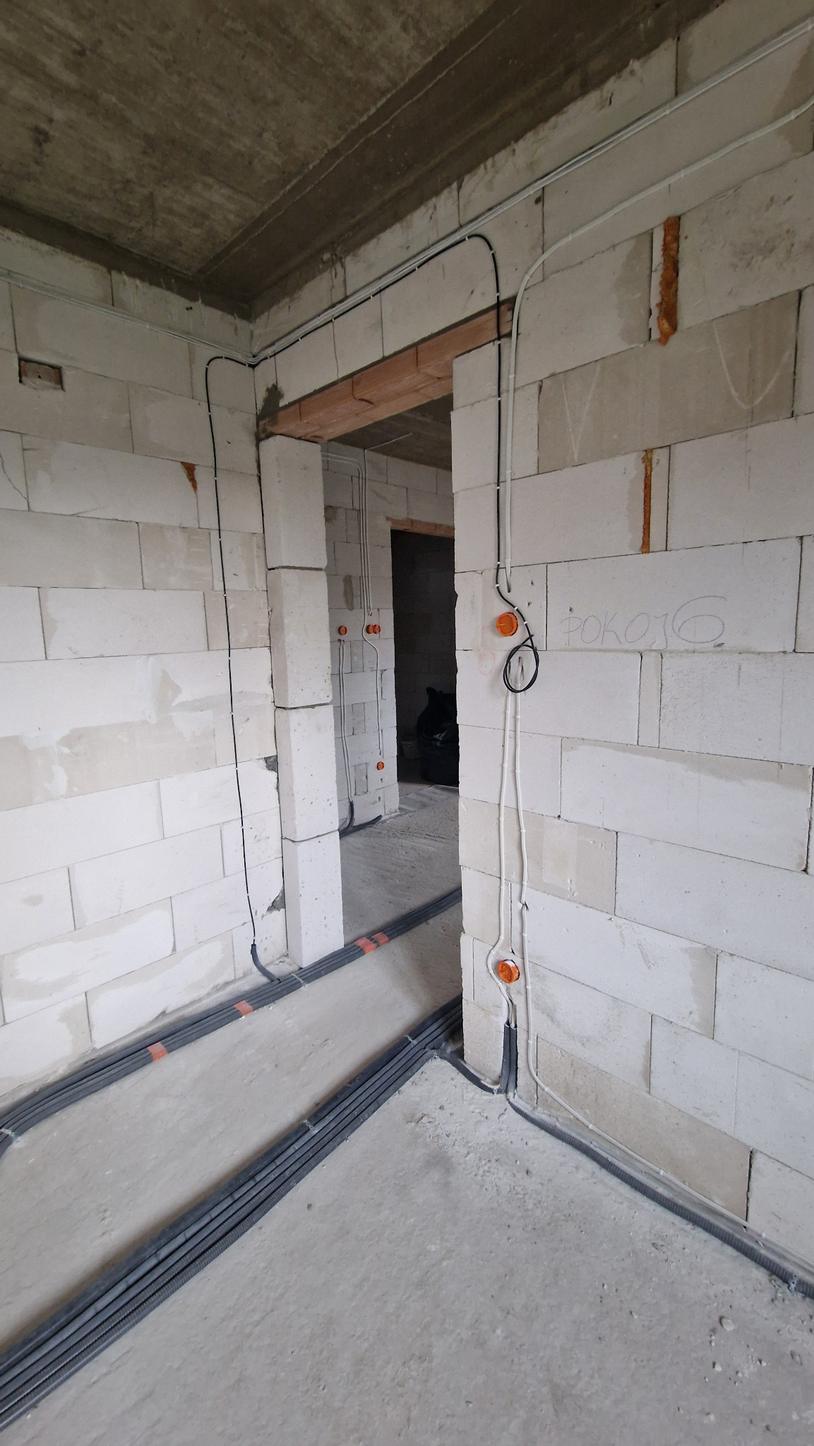 Wewnętrzne okablowanie elektryczne w surowym stanie, widoczne peszle na podłodze i przewody prowadzone po ścianach z bloczków betonowych.