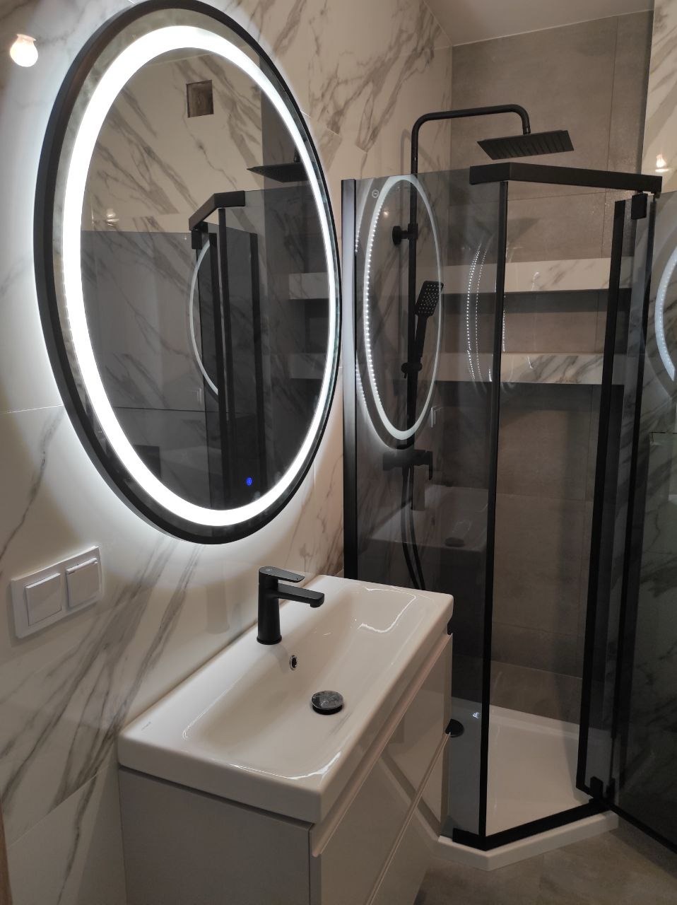 Nowoczesna łazienka z okrągłym lustrem LED, białą umywalką z czarną armaturą i kabiną prysznicową z czarnymi profilami na tle marmurowych płytek.