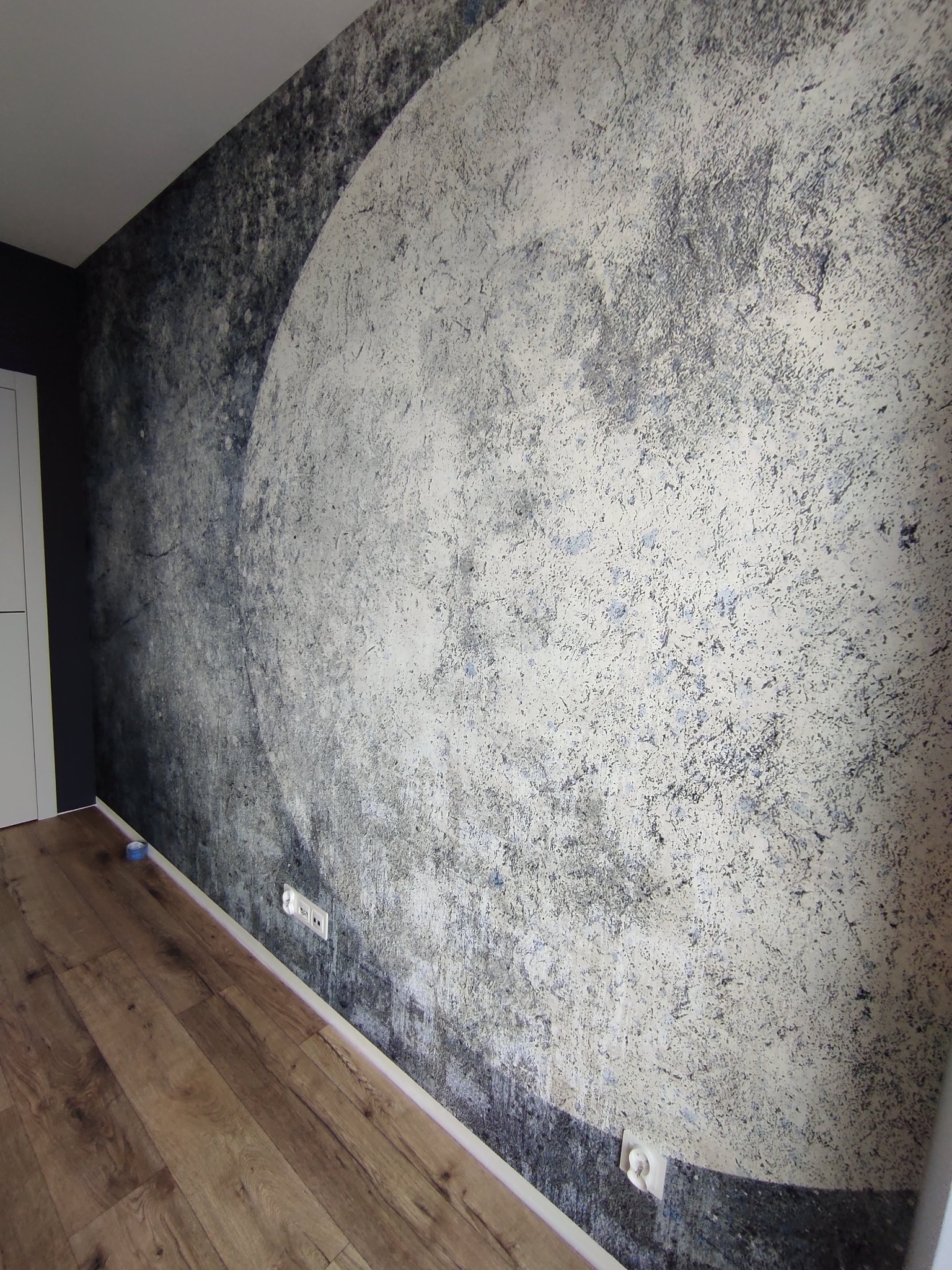 Ściana z fototapetą przedstawiającą księżyc w stylu grunge, widoczne gniazdka elektryczne i fragment podłogi z paneli.