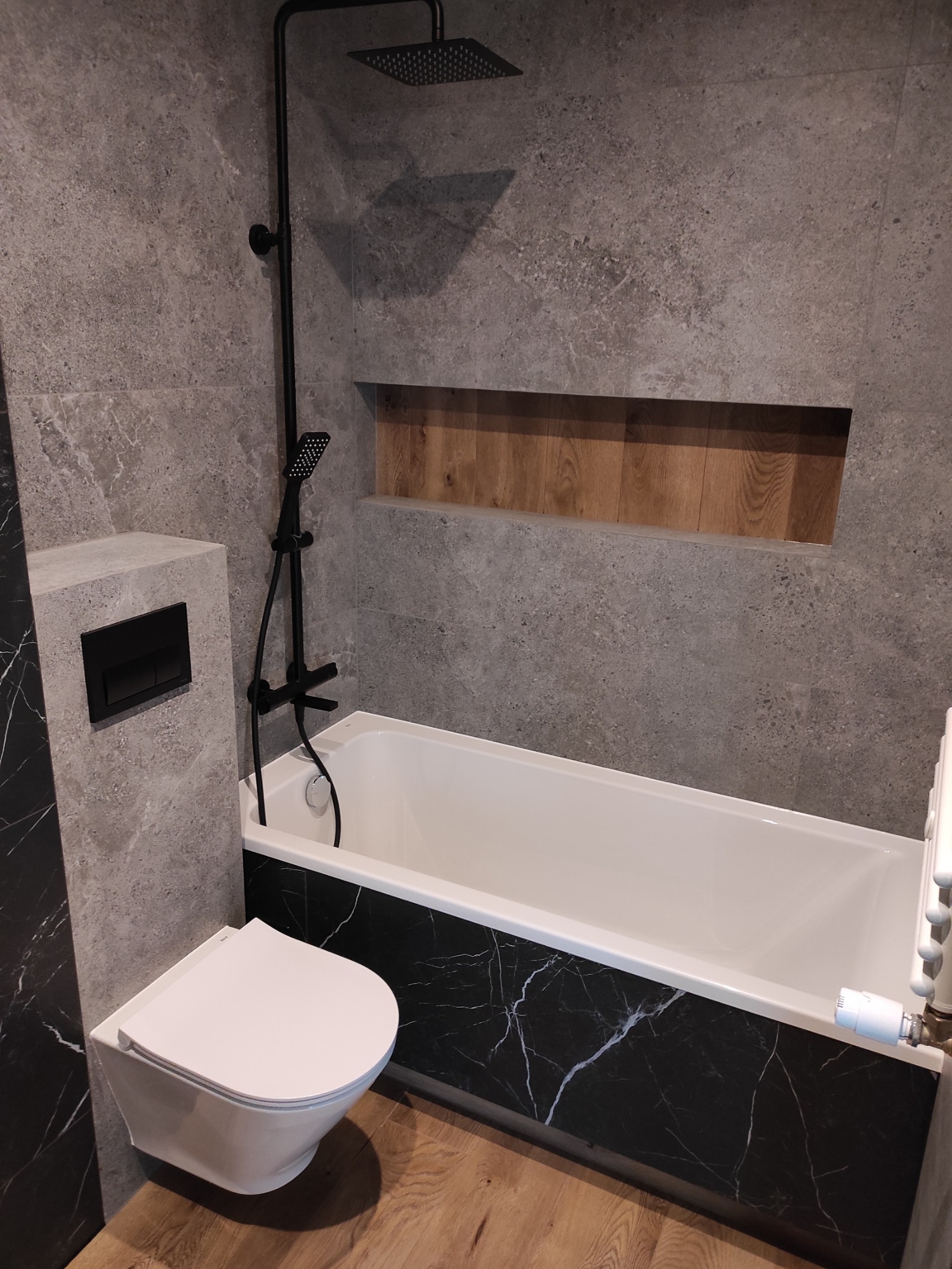 Nowoczesna łazienka z szarą płytką imitującą kamień, czarną armaturą i wanną z obudową imitującą czarny marmur, drewniana wnęka w ścianie, wisząca toaleta z czarnym przyciskiem.
