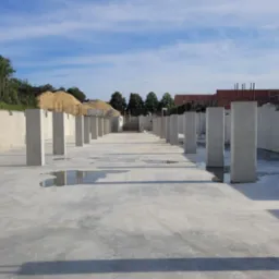 Betonowa płyta fundamentowa z pionowymi słupami, widoczne kałuże wody, w tle hałda piasku i fragment zabudowy.