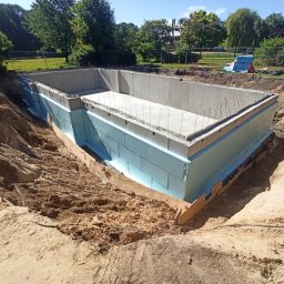 Lewandowski Constructions Sp. z o.o. - Betonowy fundament basenu z izolacją termiczną z niebieskiego styropianu, widok z góry na wykopie z ziemią, z wystającymi prętami zbrojeniowymi, w tle park z drzewami i ogrodzeniem.