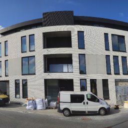 Lewandowski Constructions Sp. z o.o. - Wielopiętrowy budynek w trakcie budowy z białej cegły, widoczne okna z szarymi ramami, zaparkowane samochody dostawcze i materiały budowlane na chodniku, błękitne niebo.