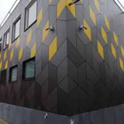 Nowoczesna elewacja budynku z szarymi i żółtymi panelami ułożonymi w geometryczny wzór rombów, widok z wąskiej uliczki miejskiej.