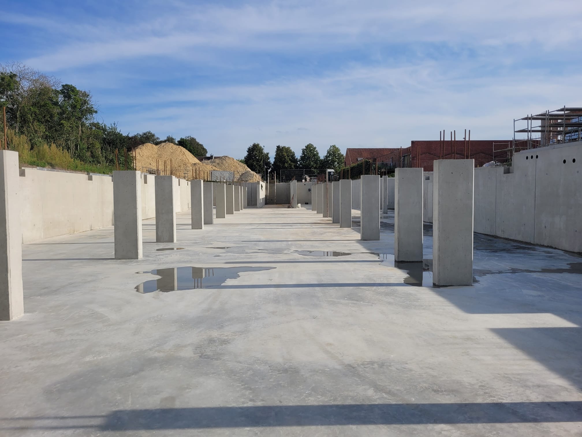 Betonowa płyta fundamentowa z pionowymi słupami, widoczne kałuże wody, w tle hałda piasku i fragment zabudowy.