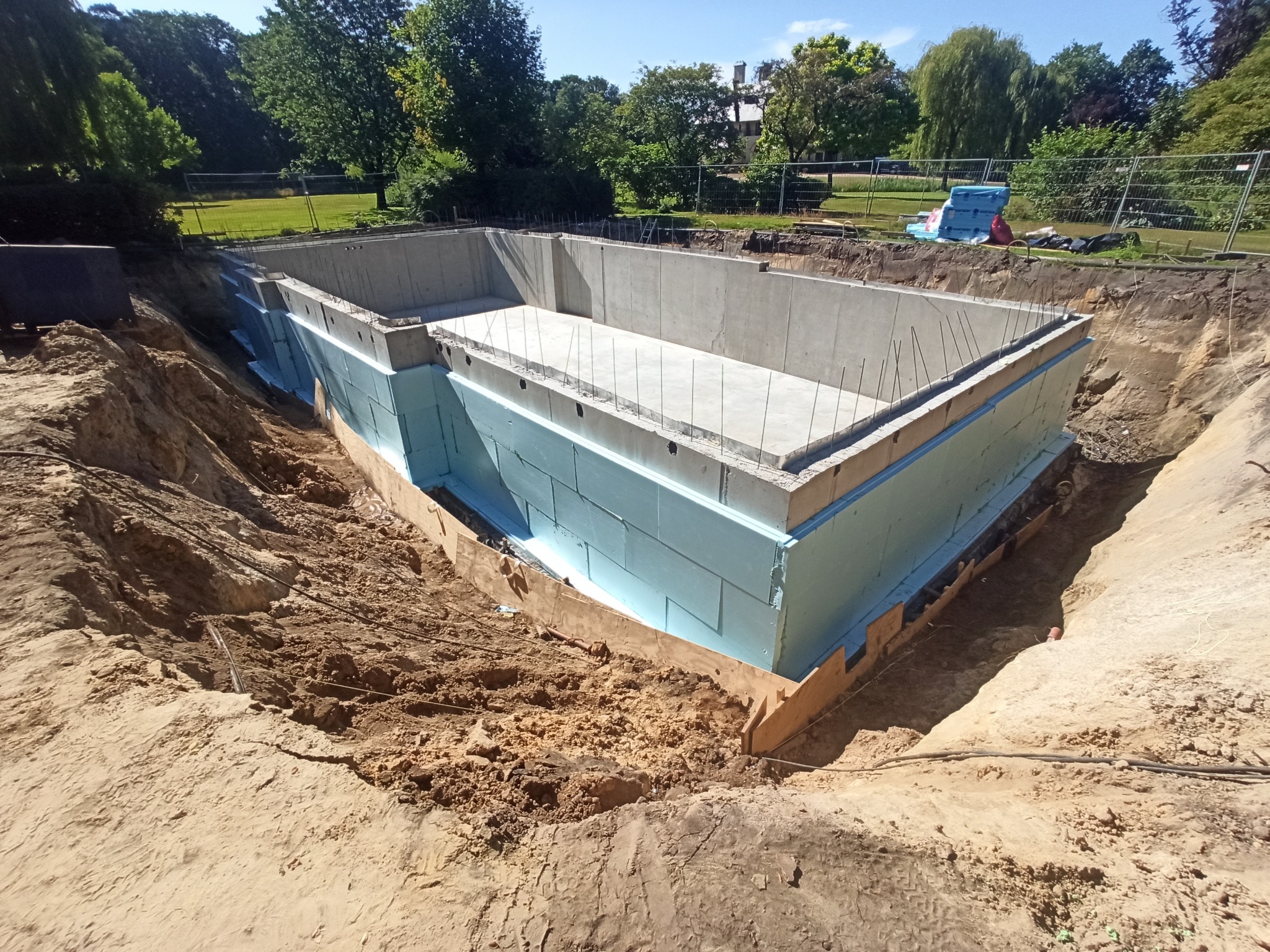 Betonowy fundament basenu z izolacją termiczną z niebieskiego styropianu, widok z góry na wykopie z ziemią, z wystającymi prętami zbrojeniowymi, w tle park z drzewami i ogrodzeniem.