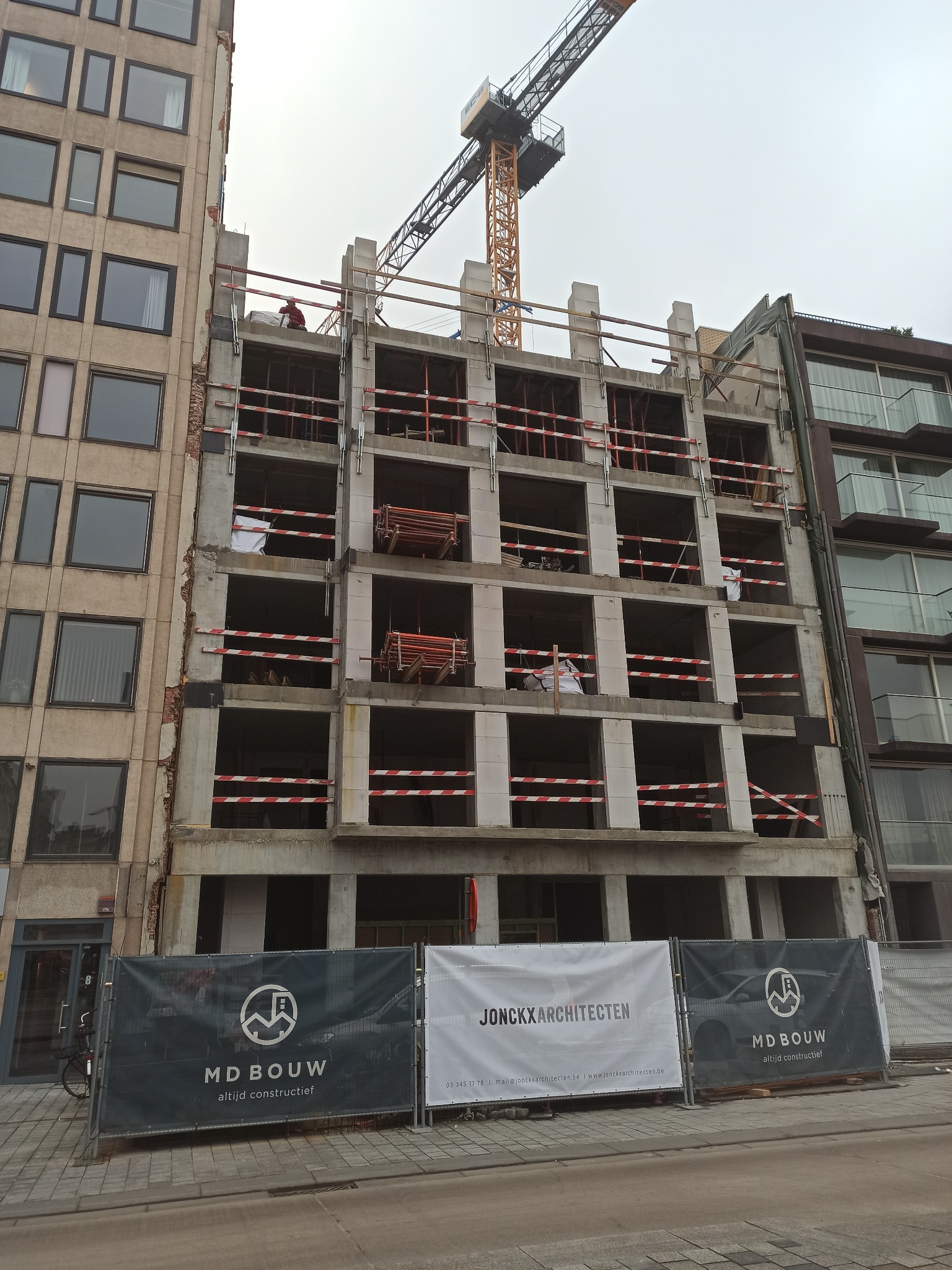 Budynek w stanie surowym z widocznym szkieletem żelbetowym, zabezpieczony taśmami ostrzegawczymi, z dźwigiem w tle i banerami firm budowlanych u dołu.