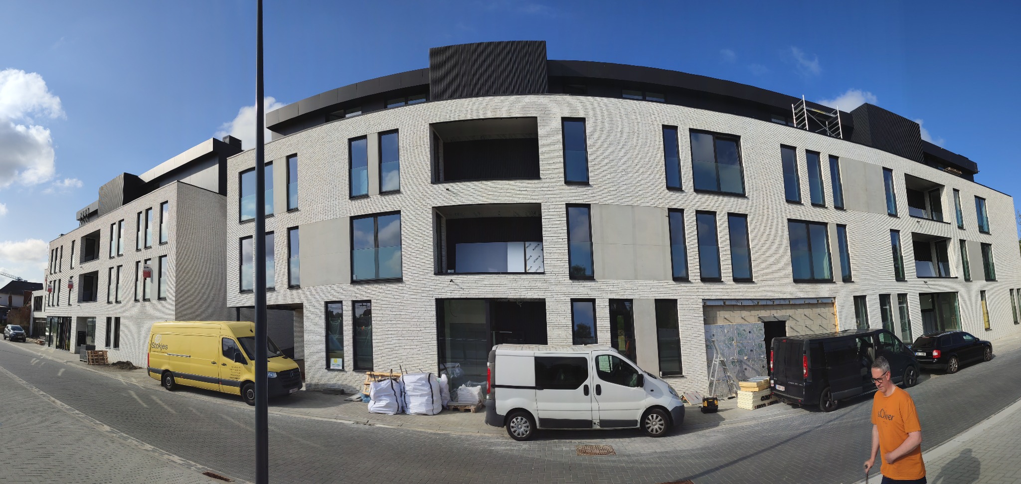 Wielopiętrowy budynek w trakcie budowy z białej cegły, widoczne okna z szarymi ramami, zaparkowane samochody dostawcze i materiały budowlane na chodniku, błękitne niebo.