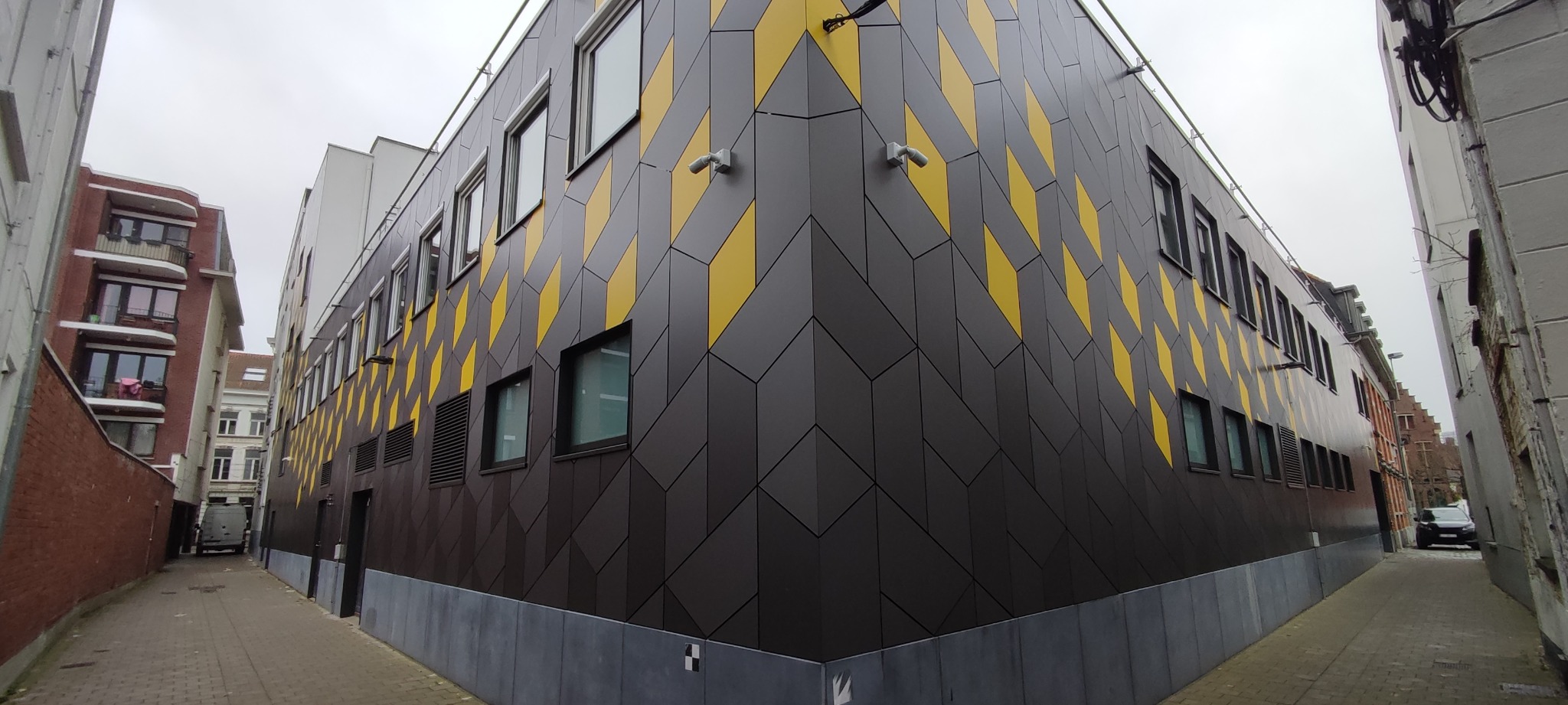 Nowoczesna elewacja budynku z szarymi i żółtymi panelami ułożonymi w geometryczny wzór rombów, widok z wąskiej uliczki miejskiej.