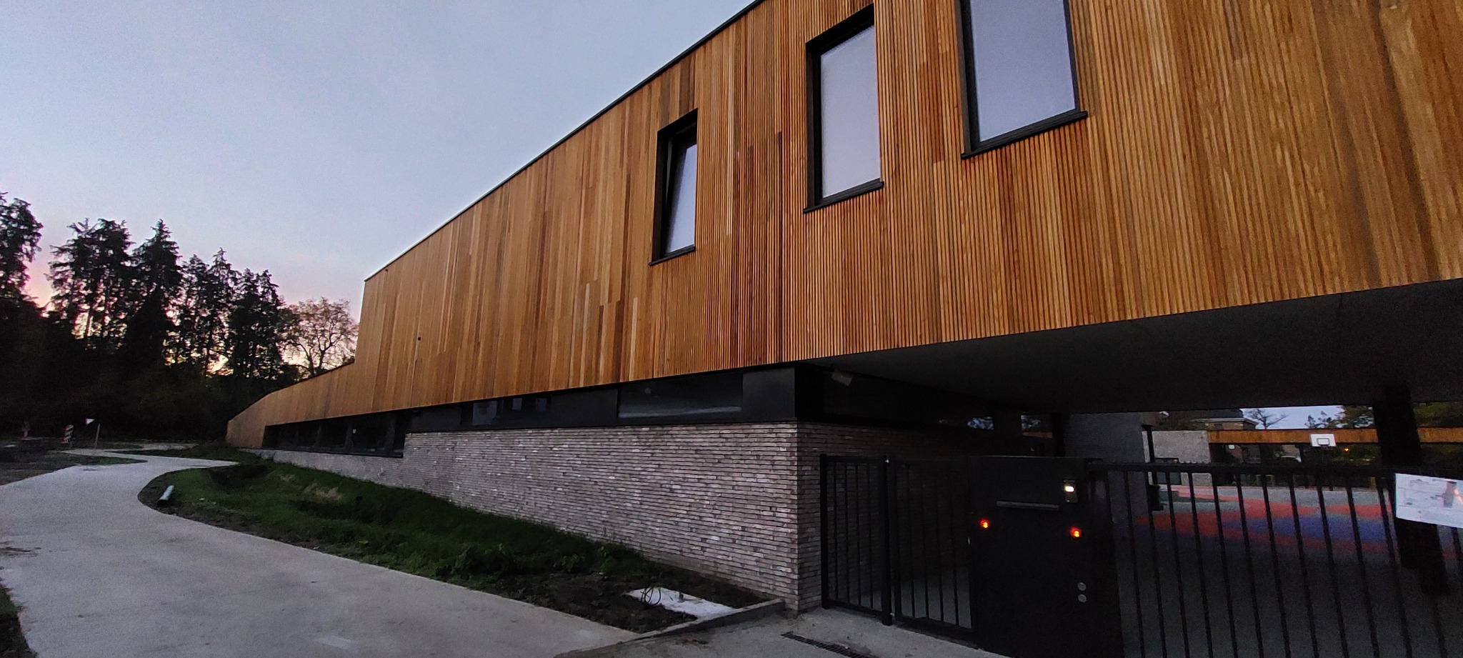 Nowoczesny dom jednorodzinny z elewacją z naturalnego drewna i cegły klinkierowej, widok z zewnątrz, brama wjazdowa z czarnego metalu, w tle drzewa i betonowy chodnik.