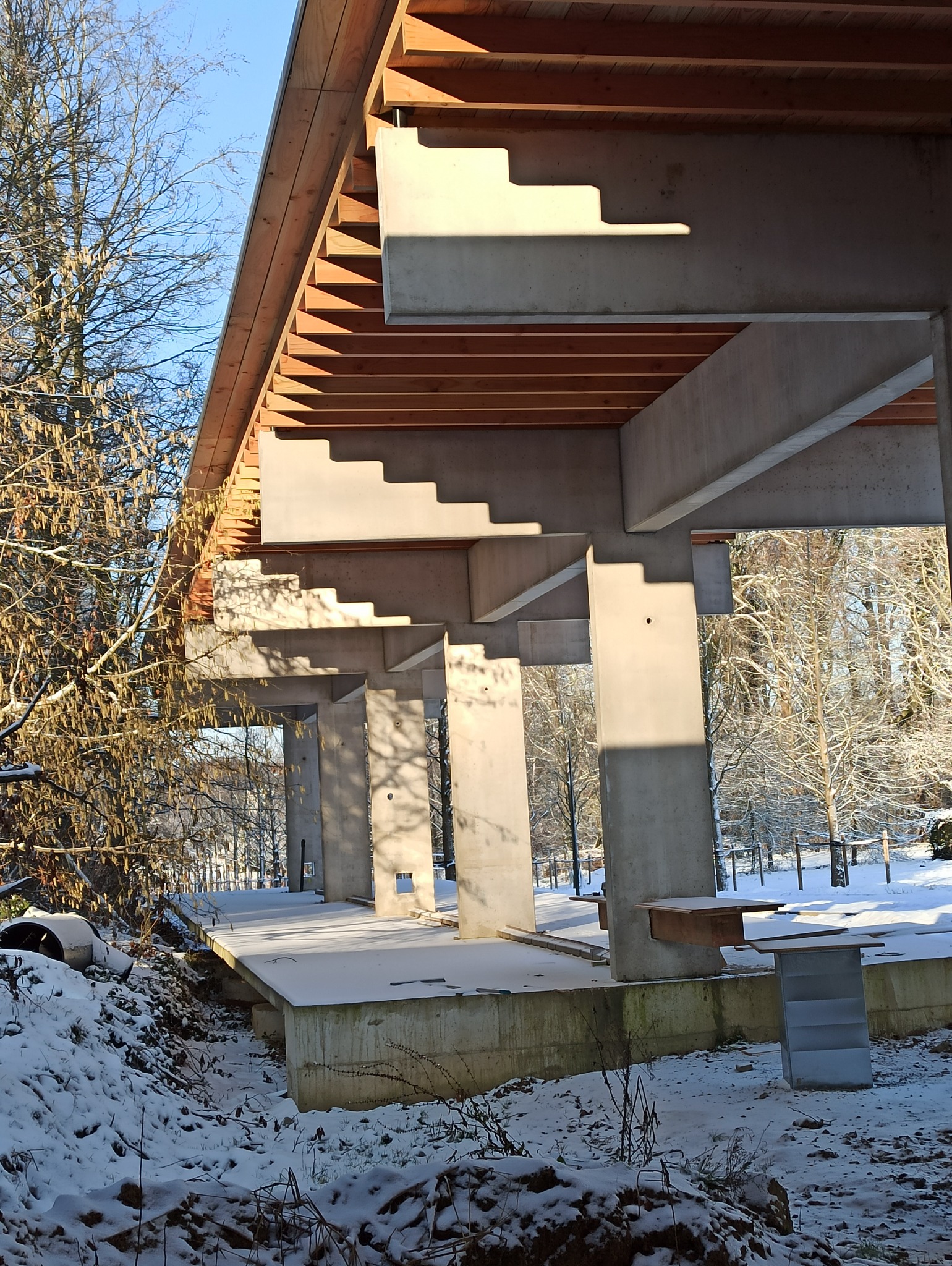 Surowa konstrukcja żelbetowa budynku w zimowej scenerii, widoczne betonowe filary i strop z drewnianym szalunkiem, pokryte śniegiem, w tle ośnieżone drzewa.