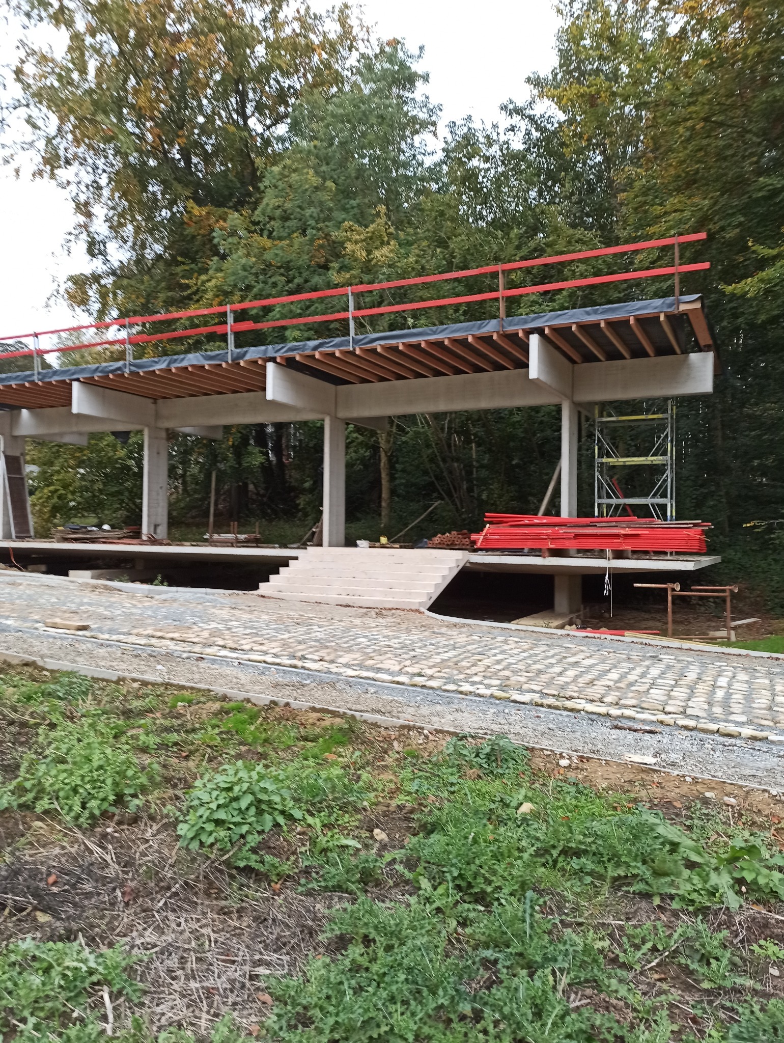 Surowa konstrukcja żelbetowa z czerwonymi barierkami ochronnymi na krawędzi, widok z poziomu gruntu na tle drzew