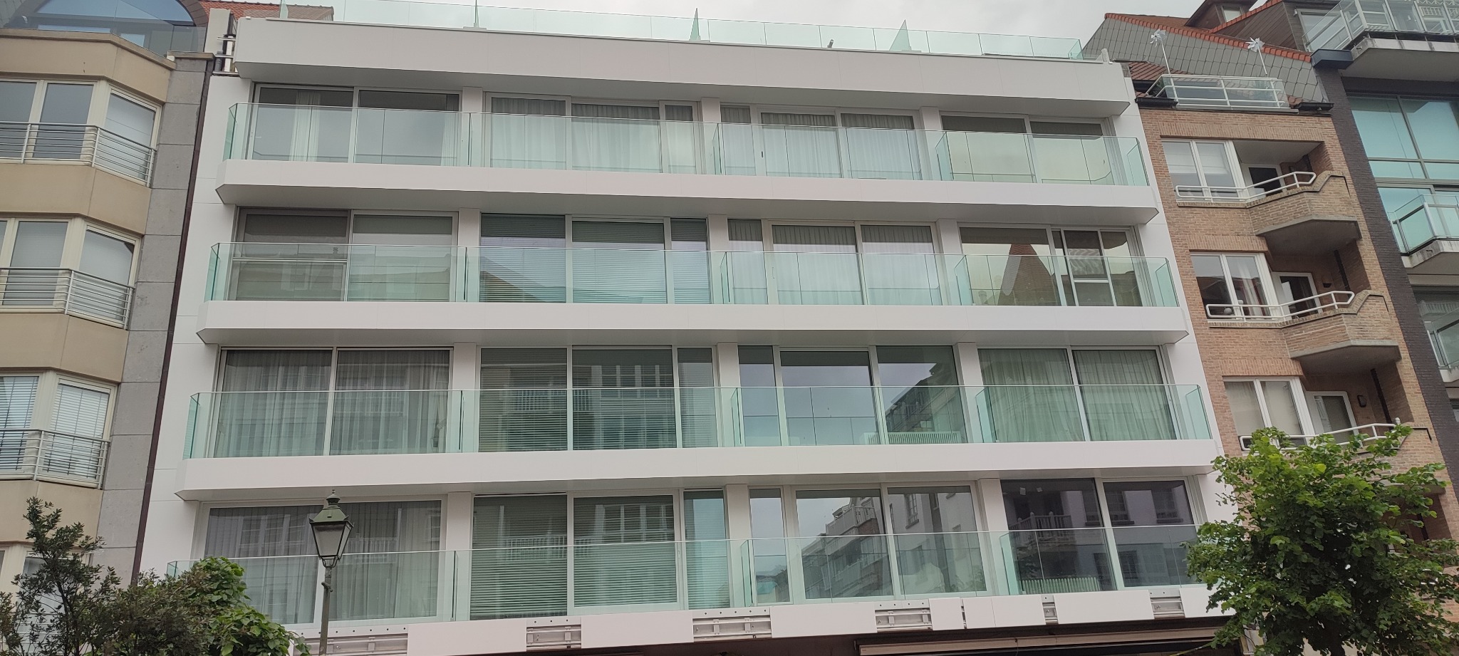 Nowoczesny, biały budynek wielorodzinny z balkonami zabezpieczonymi szkłem, widok z dołu na elewację z oknami z zasłonami lub żaluzjami.
