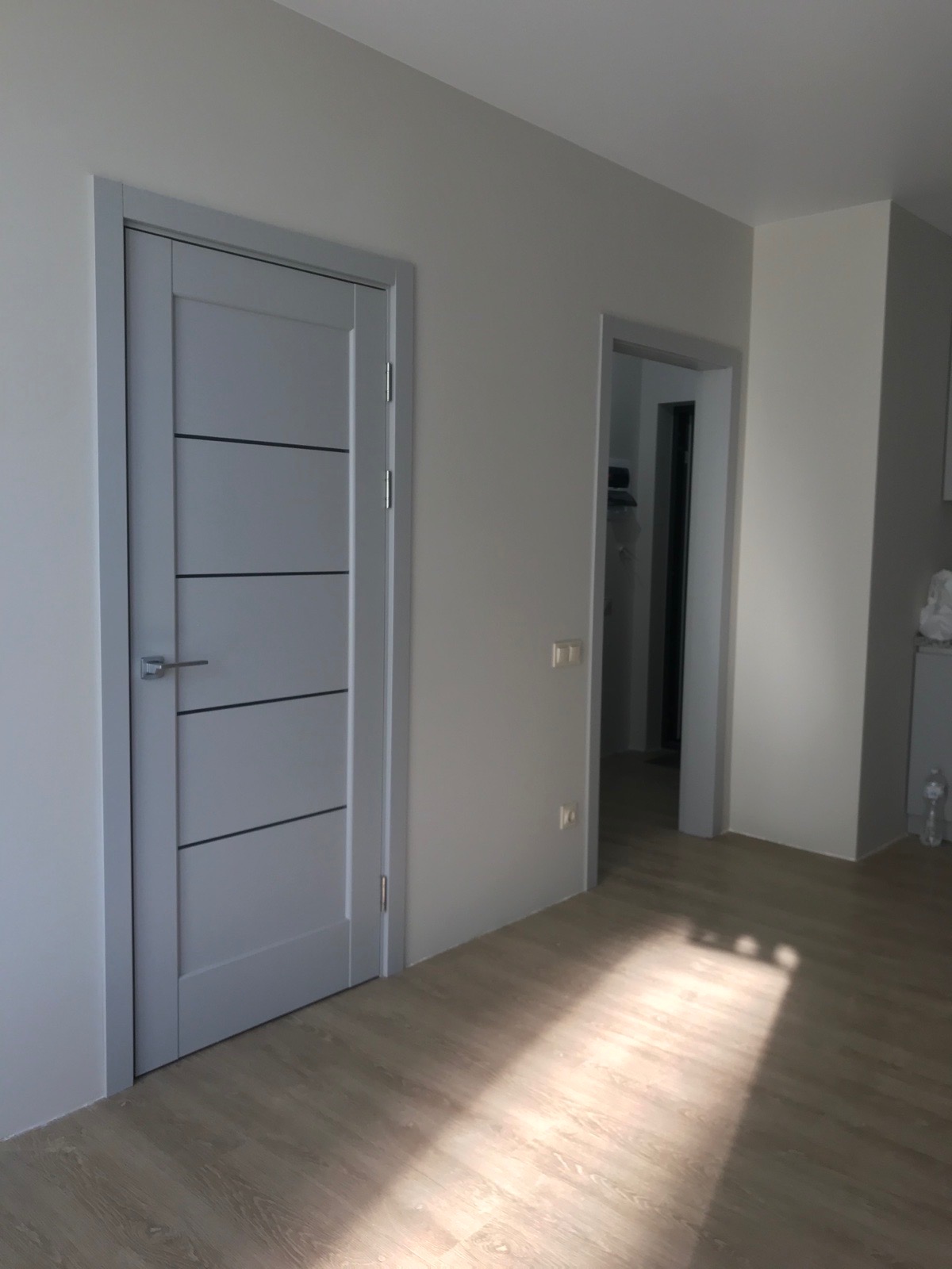 Szare drzwi wewnętrzne z poziomymi, ciemnymi paskami, zamontowane w jasnym pomieszczeniu z panelami podłogowymi, widok na otwarte drzwi do garderoby w tle.