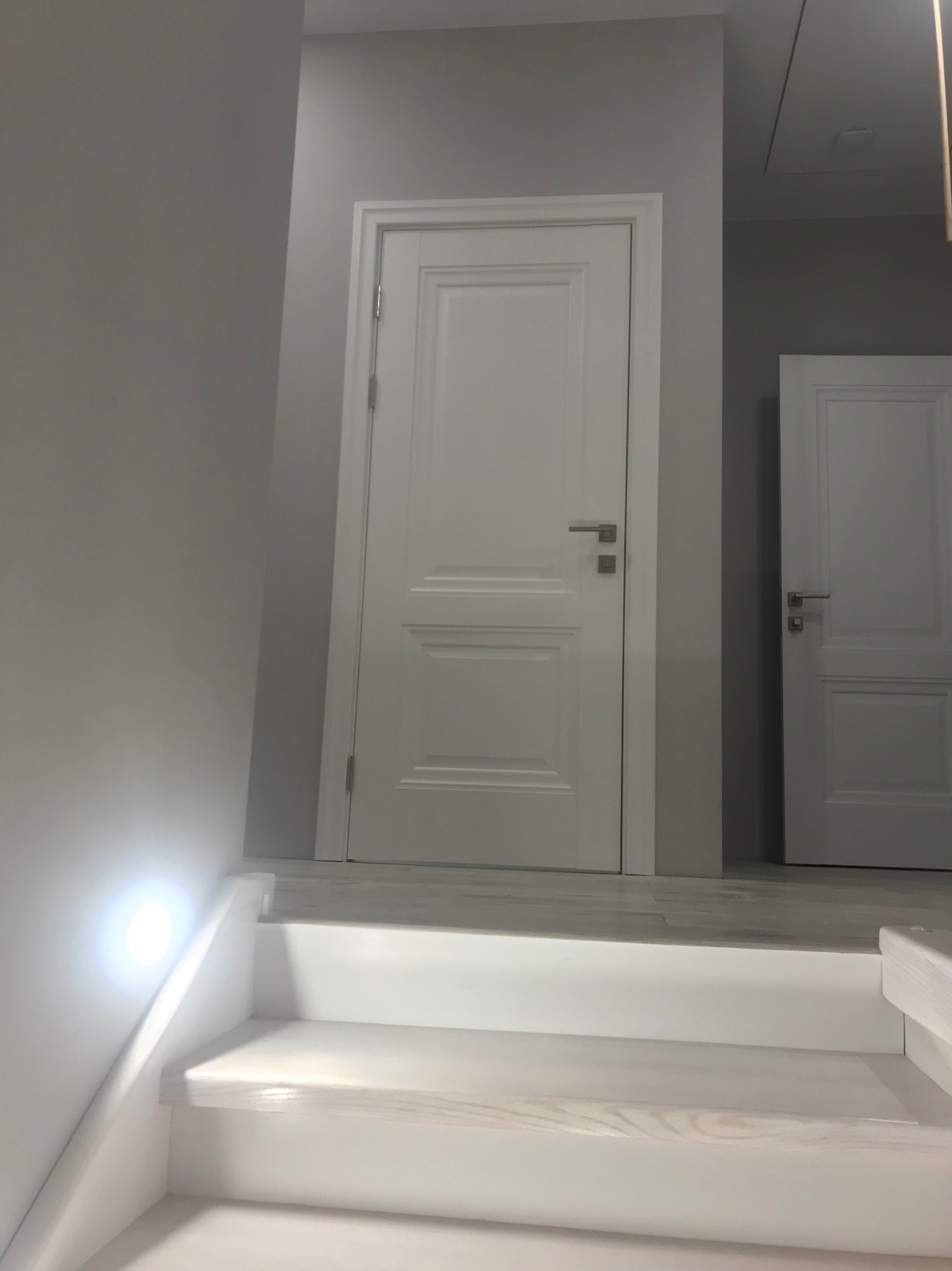 Białe, panelowe drzwi wewnętrzne z srebrną klamką, widoczne ze schodów w jasnym, minimalistycznym wnętrzu. W tle drugie, podobne drzwi.