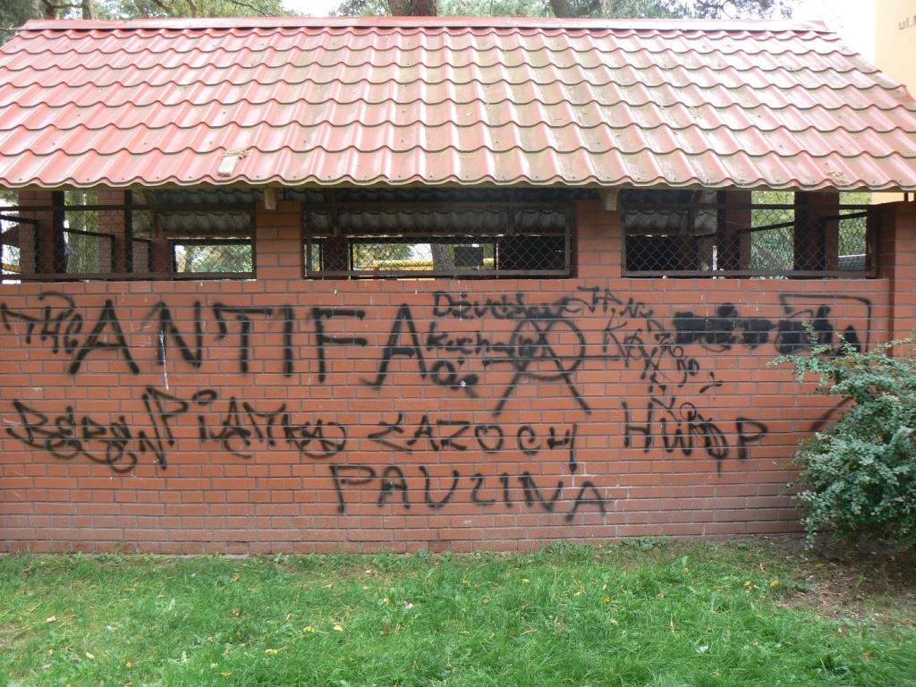 Ceglana ściana budynku z czerwonym dachem pokryta czarnymi graffiti, widoczne napisy 'Antifa' i inne, zarośnięta trawa u podstawy.