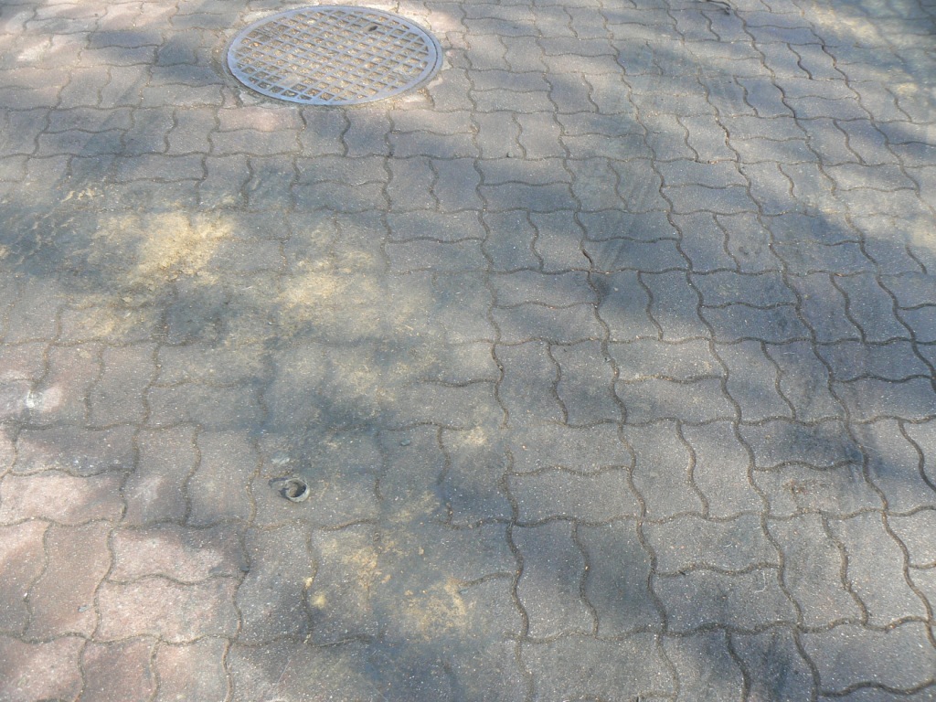 Szara kostka brukowa po czyszczeniu, widoczne ślady po plamach i okrągła studzienka kanalizacyjna w rogu. Częściowo zacieniona powierzchnia.