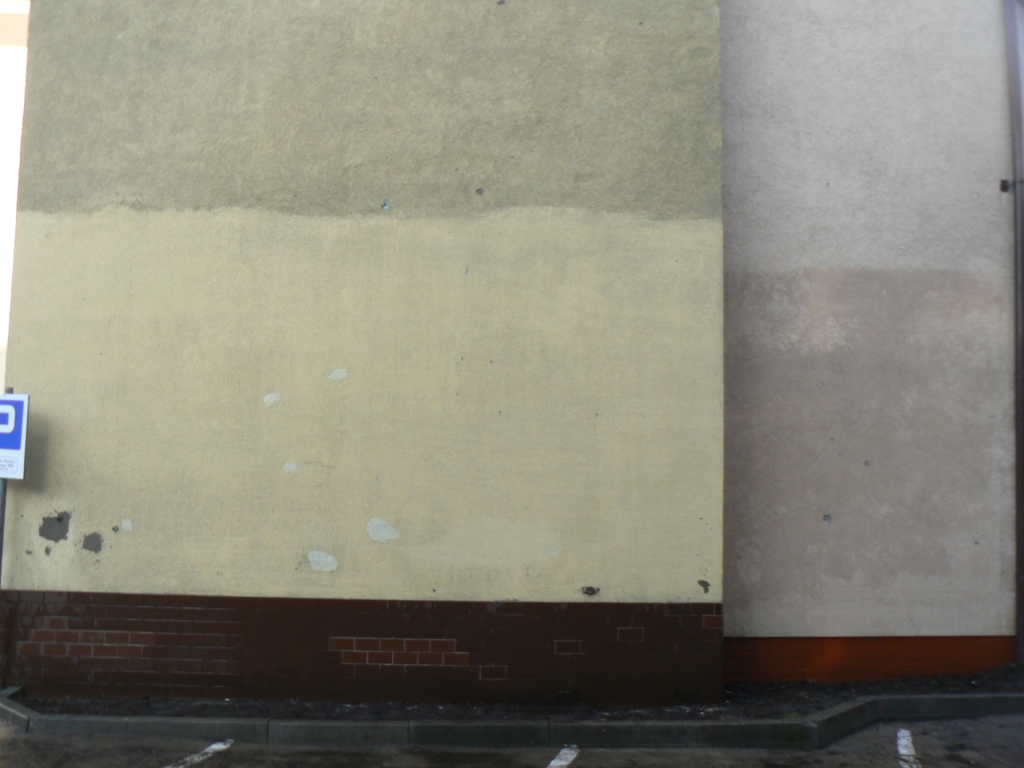 Fragment ściany budynku z widocznymi śladami zabrudzeń i przebarwień przed czyszczeniem, z ciemną, ceglaną podmurówką i znakiem parkingu po lewej stronie.