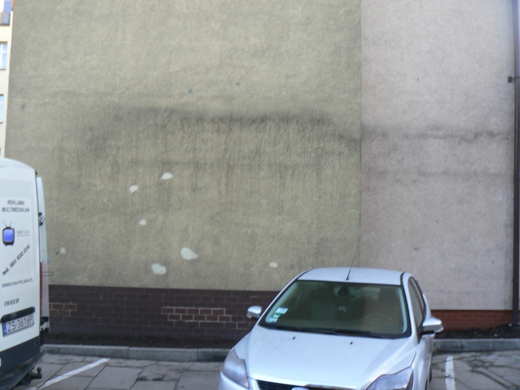 Brudna elewacja budynku z widocznymi śladami po czyszczeniu miejscowym, zaparkowany srebrny samochód osobowy i biały bus z reklamą. Częściowo oczyszczona powierzchnia muru kontrastuje z zabrudzoną.