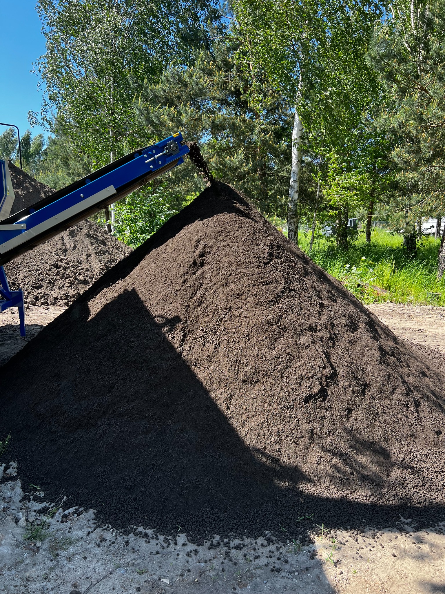 Transport ciężarowy w Białymstoku: taśmociąg podający ziemię na pryzmę, widoczne drzewa w tle.
