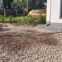 Edom.company - Instalacja drenażu rozsączającego wokół domu, widoczna rura pionowa z rozpryskiwaczem nawadniającym, otoczona kamieniami, w tle budynek i zaparkowany żółty samochód.