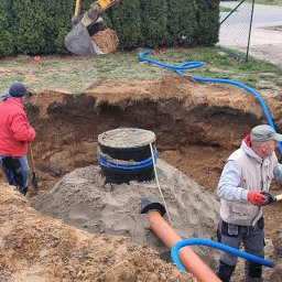 Edom.company - Montaż przydomowej oczyszczalni ścieków: dwóch pracowników w wykopie, jeden z łopatą w czerwonej kurtce, drugi z łopatą w kamizelce, widoczny zbiornik, rury i koparka w tle.
