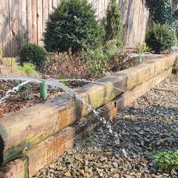 Edom.company - Drenaż liniowy z drewnianych bali w ogrodzie, z widocznymi dyszami rozpryskującymi wodę, otoczony kamieniami i roślinnością.