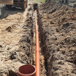 Edom.company - Pomarańczowa rura kanalizacyjna ułożona w wykopie, z widocznym operatorem koparki w tle, podczas instalacji na działce.