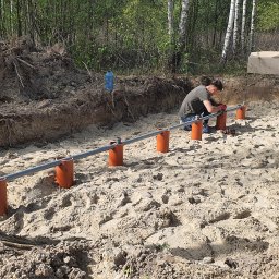 Edom.company - Montaż rur drenarskich w wykopie wypełnionym piaskiem, widoczny pracownik w rękawicach i niebieskich butach, w tle las brzozowy i wykopana ziemia.