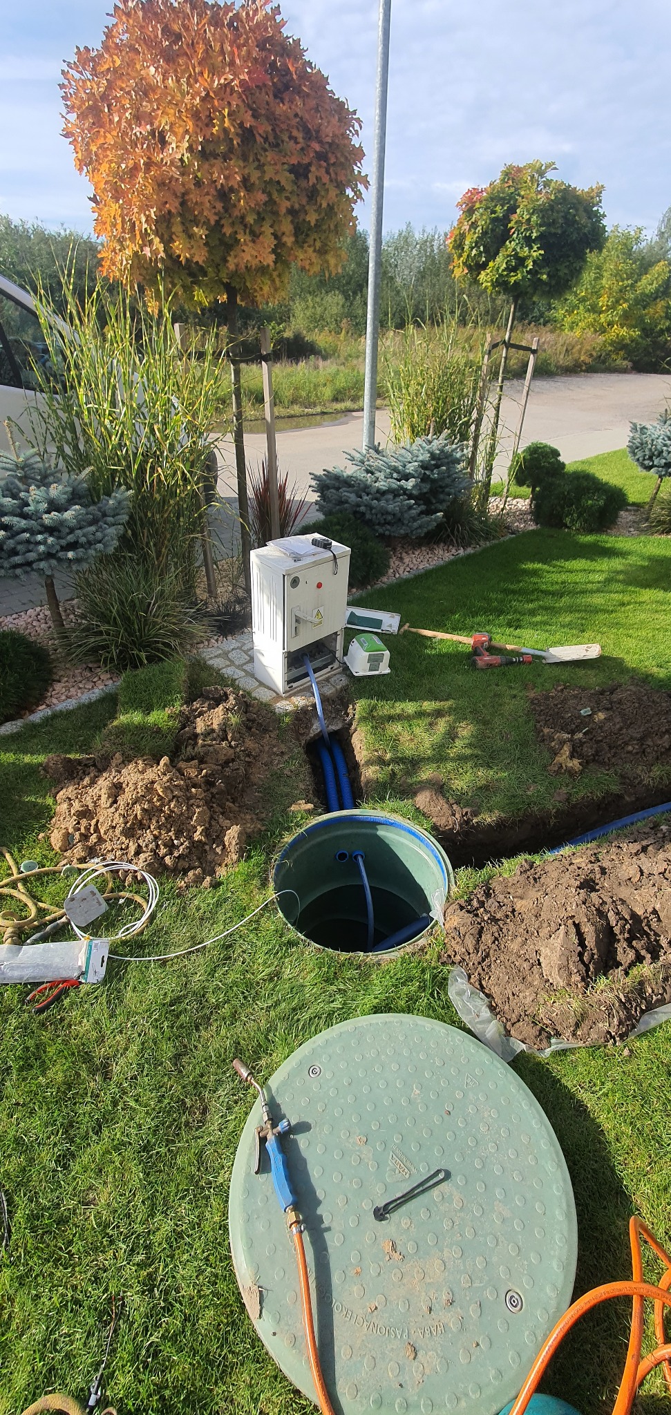 Montaż zbiornika z zieloną pokrywą w wykopie na trawniku, widoczne niebieskie rury, skrzynka elektryczna i narzędzia, w tle ozdobne drzewa i krzewy.
