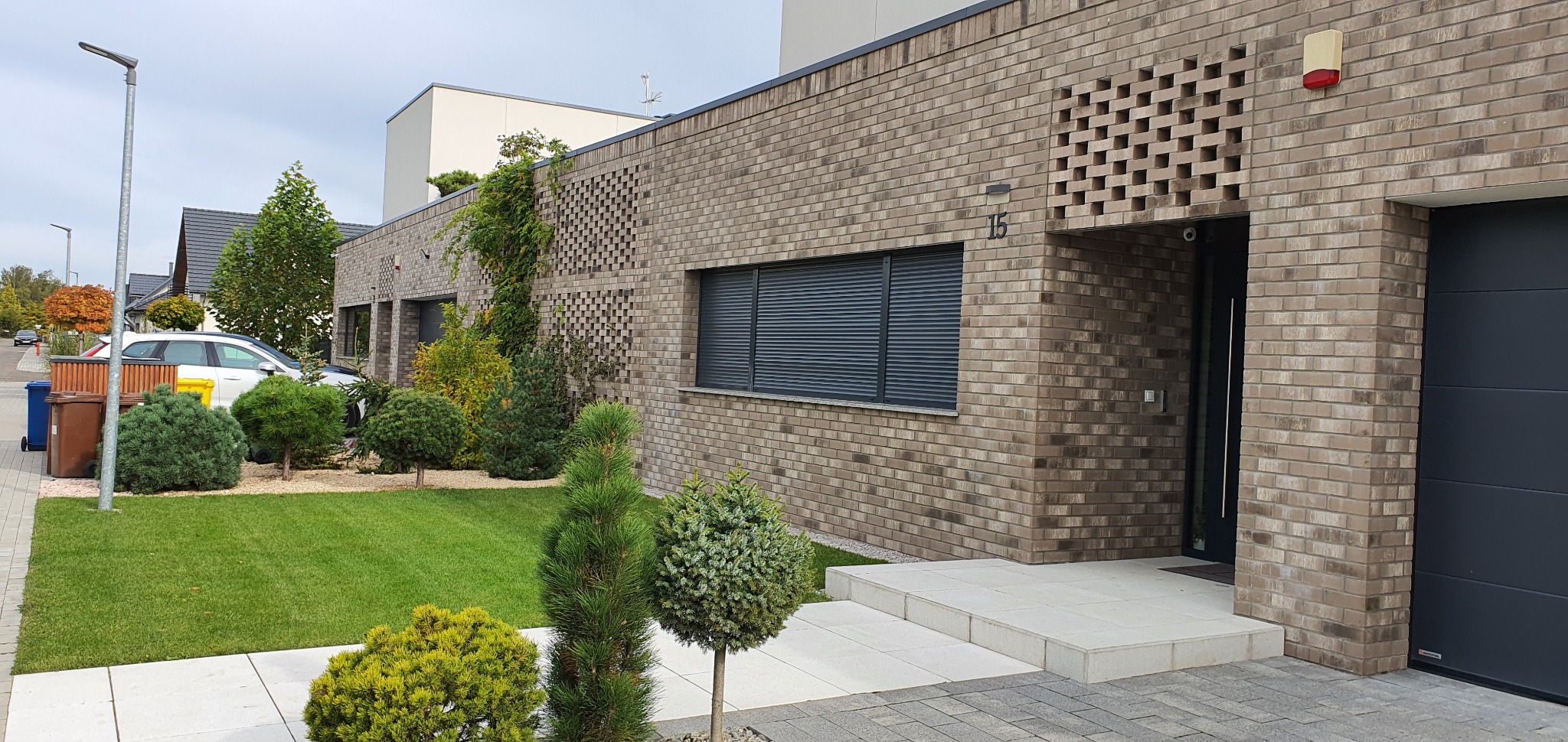 Nowoczesny dom z cegły klinkierowej z elementami ażurowymi, ciemnymi roletami i szarą bramą garażową, otoczony zadbanym trawnikiem i ozdobnymi krzewami