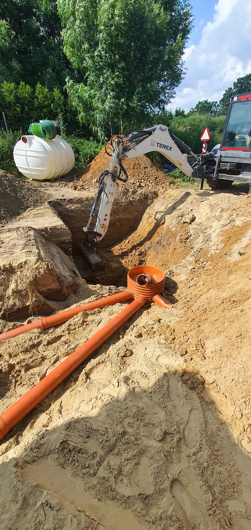 Wykonanie wykopu pod instalację systemu odprowadzania ścieków: biały zbiornik, pomarańczowe rury kanalizacyjne i koparka Terex w wykopie z piaskiem.