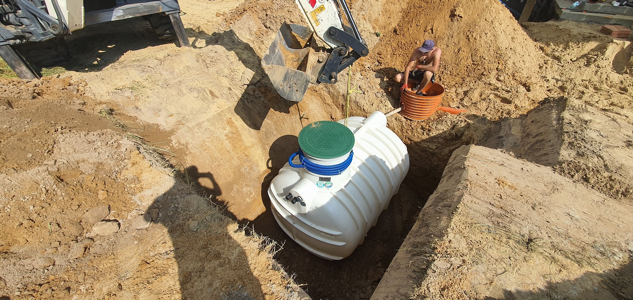 Biały zbiornik z zieloną pokrywą w wykopie, obok pomarańczowa studzienka rewizyjna i koparka, widoczny mężczyzna w niebieskiej czapce wewnątrz studzienki.