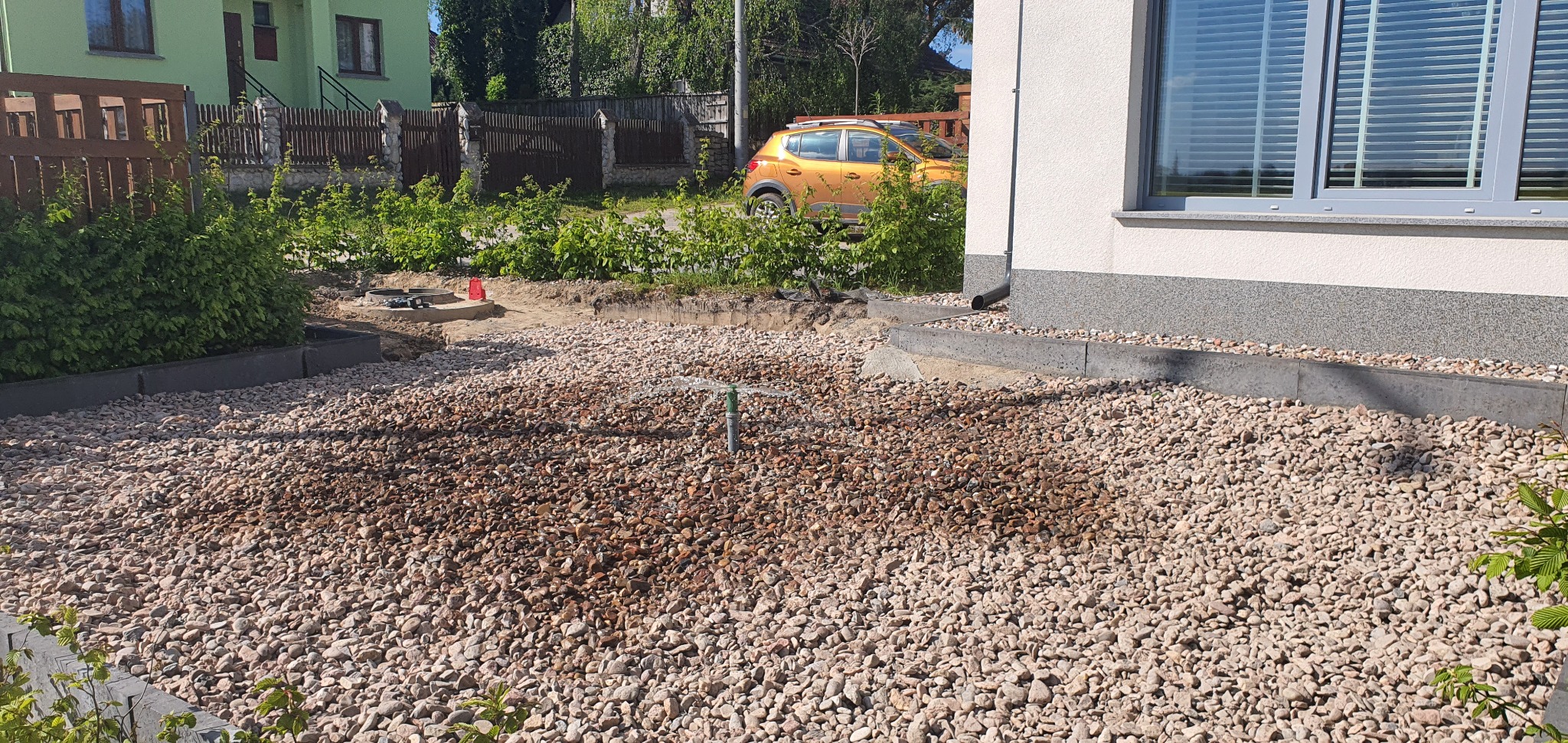 Instalacja drenażu rozsączającego wokół domu, widoczna rura pionowa z rozpryskiwaczem nawadniającym, otoczona kamieniami, w tle budynek i zaparkowany żółty samochód.