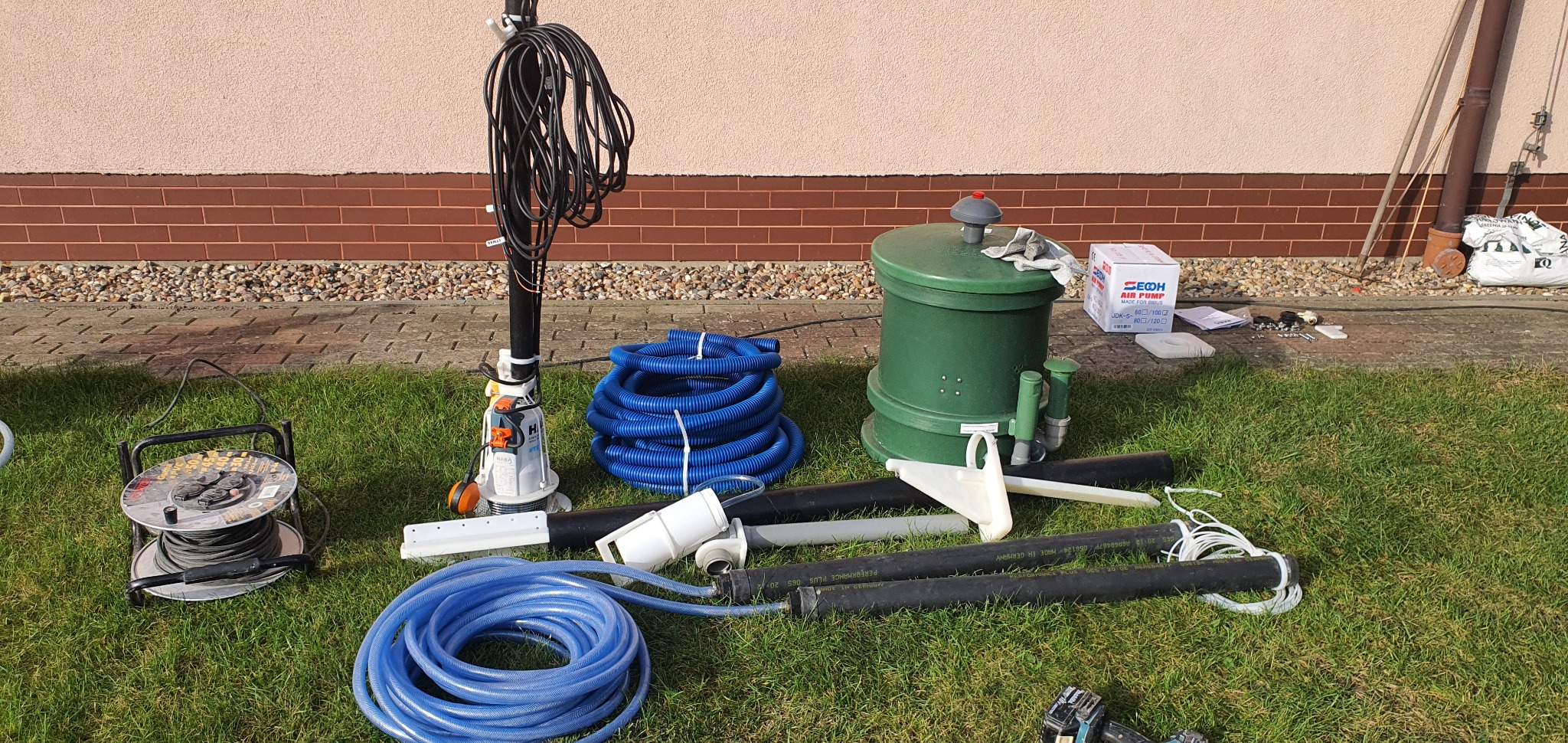 Elementy instalacji przydomowej oczyszczalni ścieków rozłożone na trawie przed budynkiem, widoczna pompa zanurzeniowa, rury, przewody, złączki oraz zielony zbiornik i karton z napisem 'AIR PUMP'.
