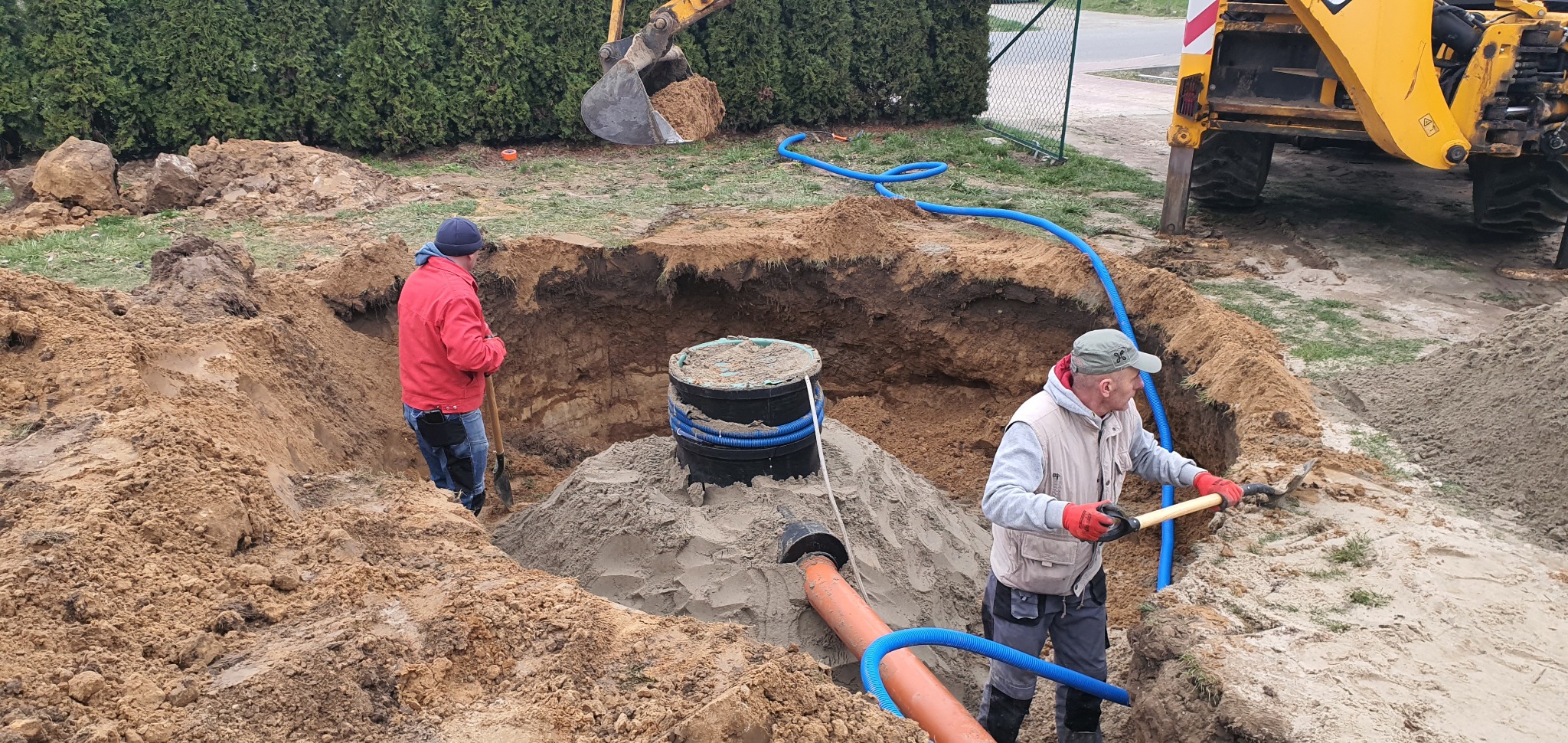 Montaż przydomowej oczyszczalni ścieków: dwóch pracowników w wykopie, jeden z łopatą w czerwonej kurtce, drugi z łopatą w kamizelce, widoczny zbiornik, rury i koparka w tle.