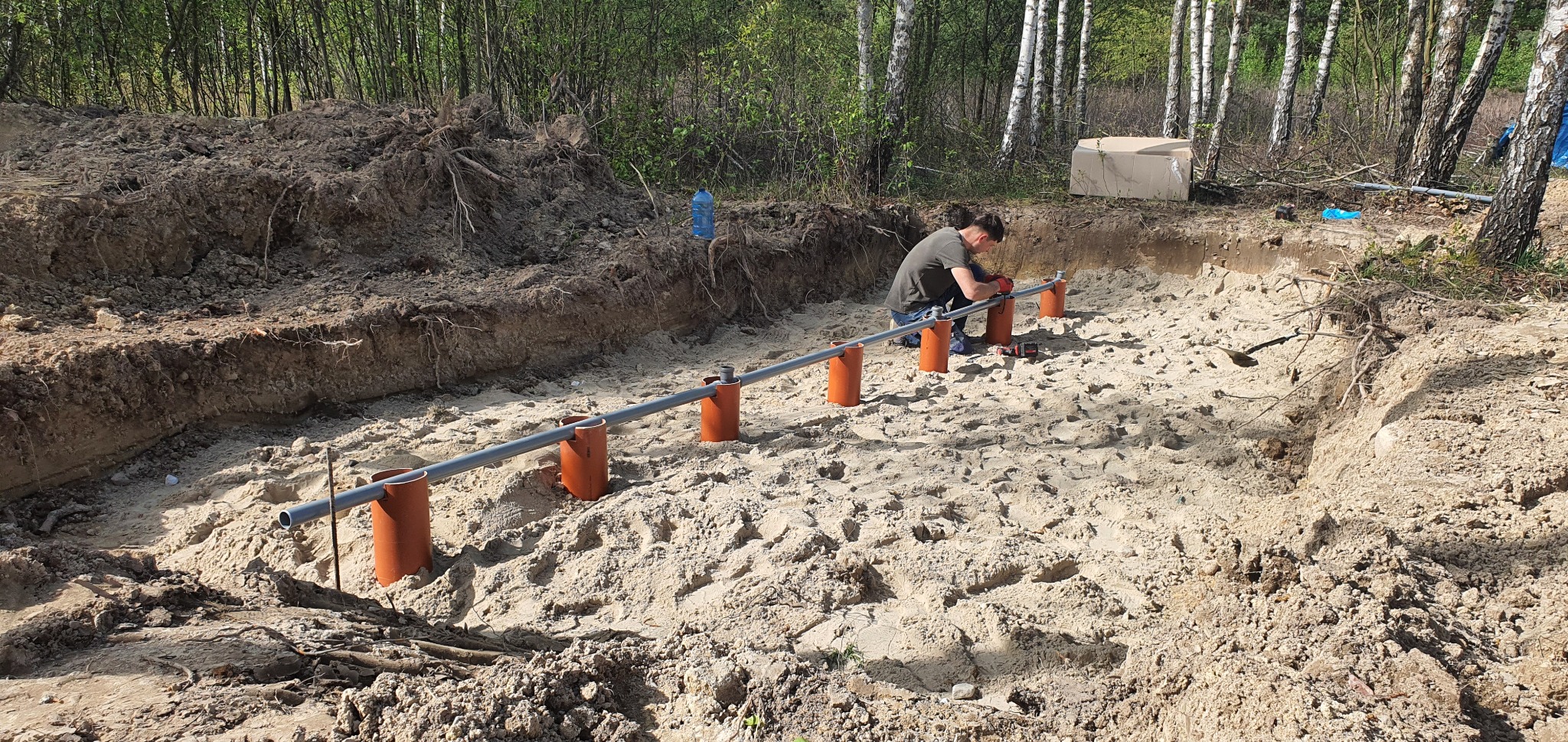 Montaż rur drenarskich w wykopie wypełnionym piaskiem, widoczny pracownik w rękawicach i niebieskich butach, w tle las brzozowy i wykopana ziemia.