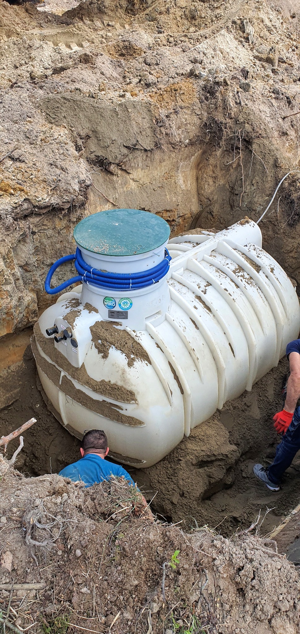 Biały zbiornik z zieloną pokrywą, prawdopodobnie osadnik wstępny, umieszczony w wykopie ziemnym, z widocznymi niebieskimi rurami karbowanymi i fragmentami sylwetek dwóch osób.
