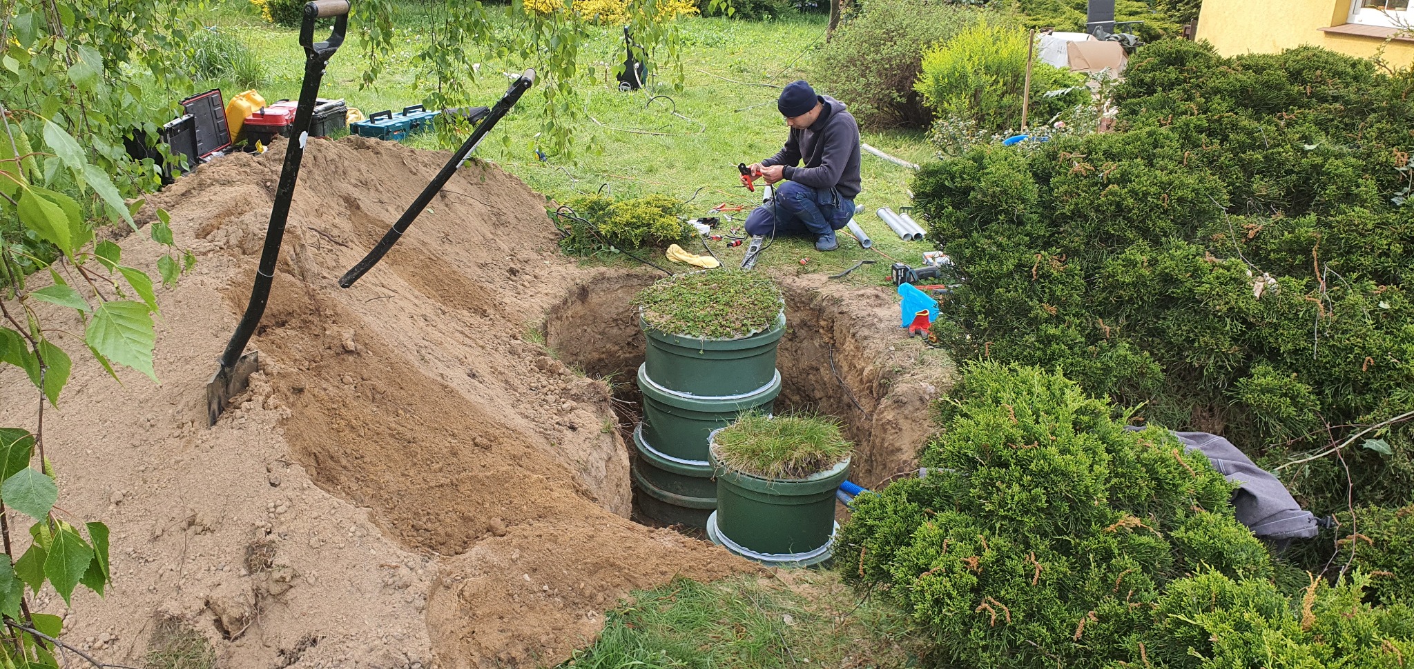 Instalacja zielonej, trzypoziomowej, przydomowej oczyszczalni ścieków w wykopie ziemnym, z widocznymi narzędziami i pracownikiem przygotowującym rury.