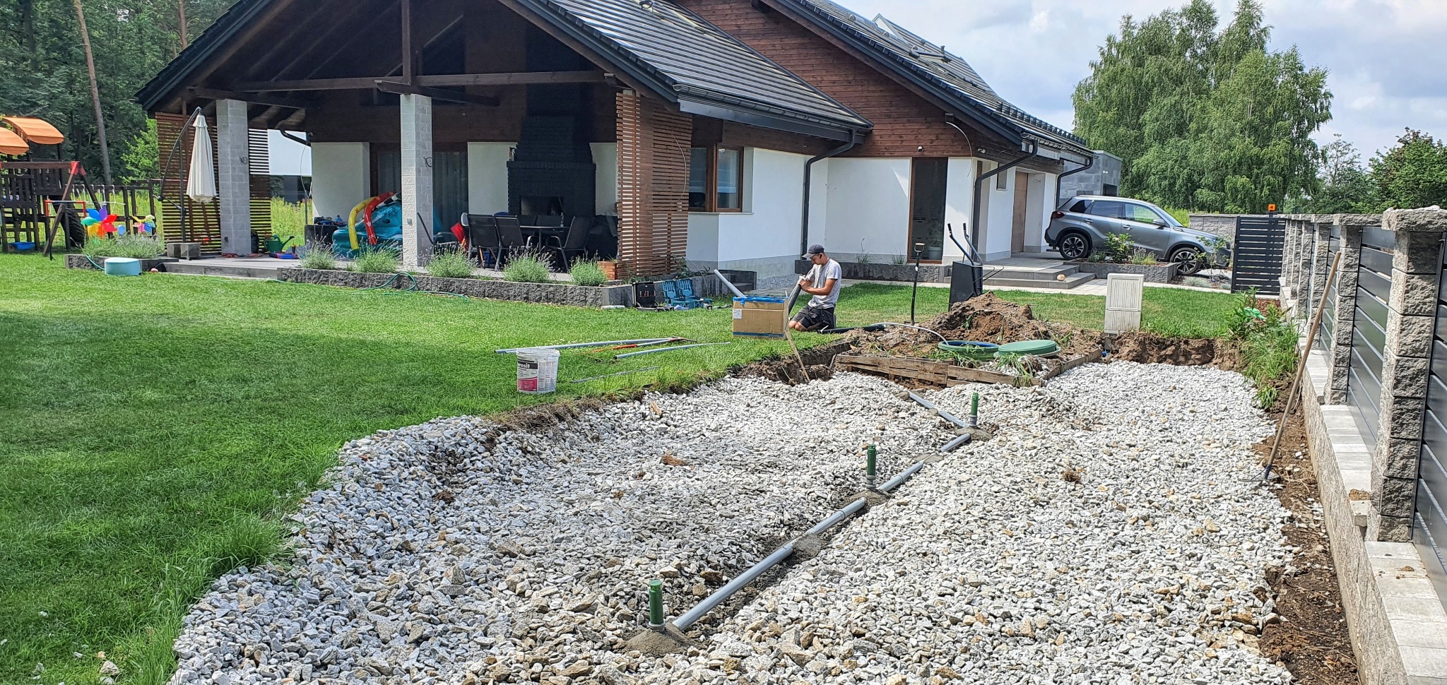 Budowa drenażu rozsączającego przydomowej oczyszczalni ścieków na działce z widocznym domem jednorodzinnym, rurami drenażowymi i warstwą żwiru; instalator w trakcie pracy łączy elementy systemu.
